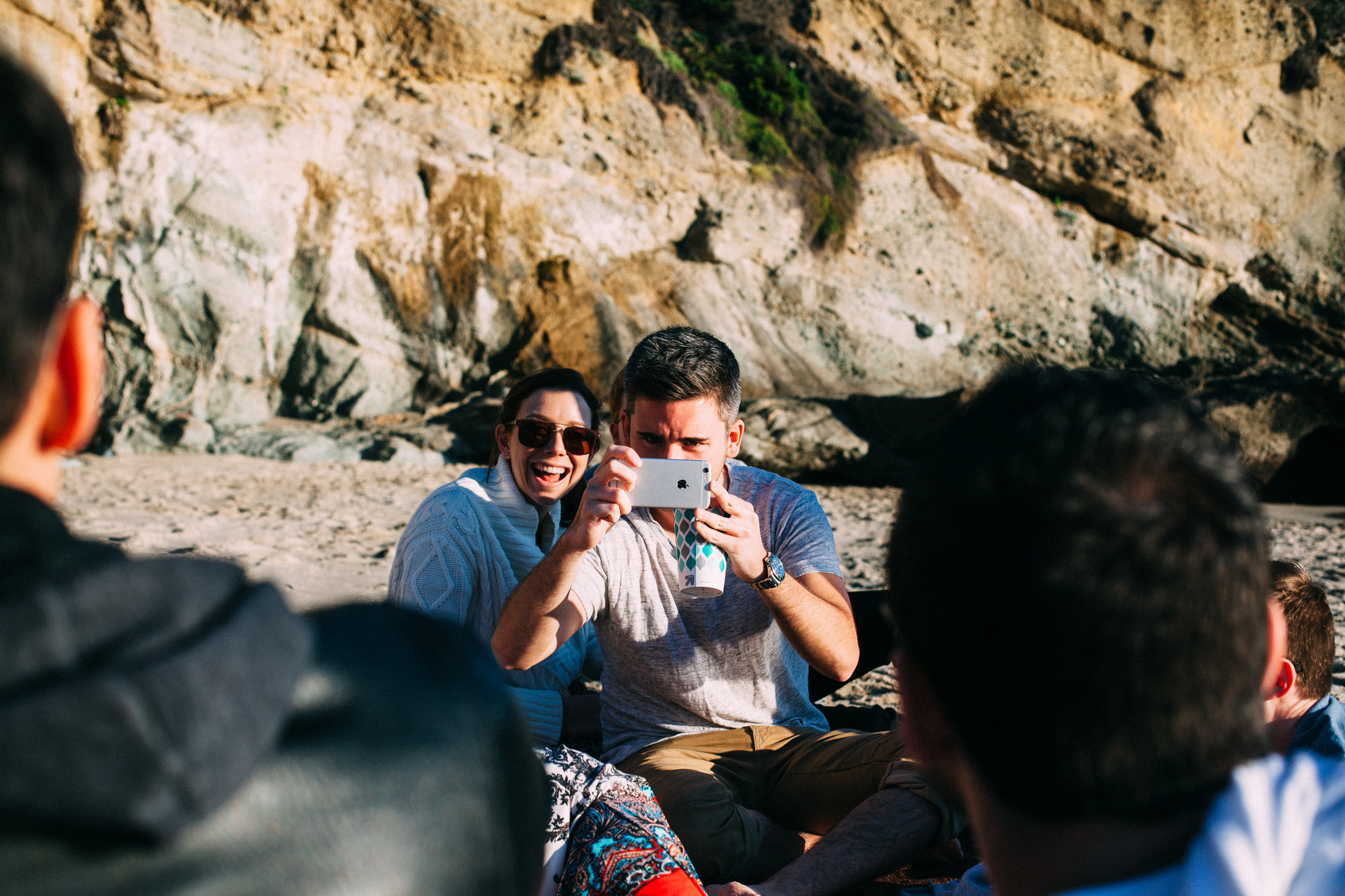Young man holding up a phone beside a laughing woman in sunglasses and a cream knit sweater on the beach.