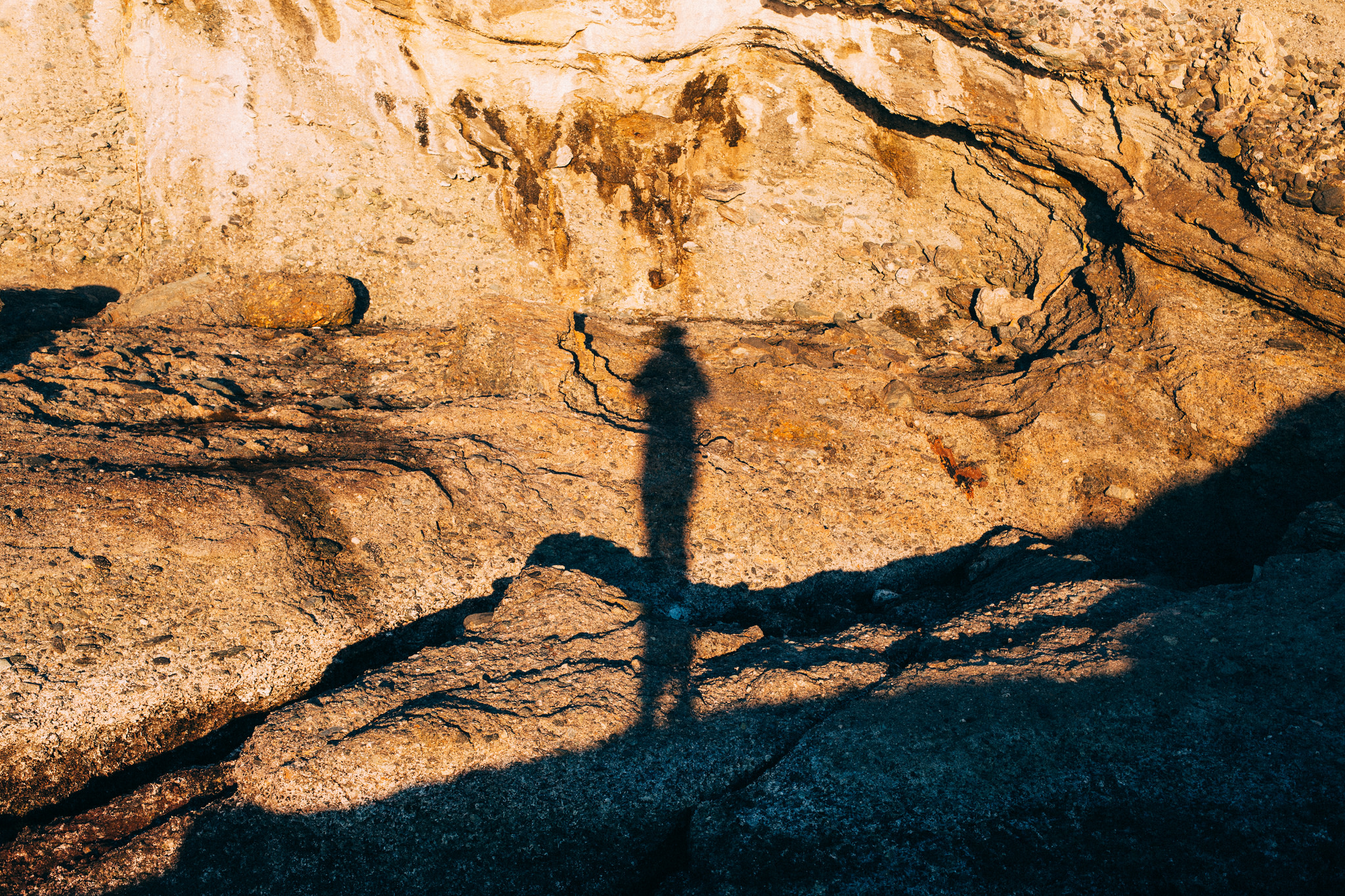 Long shadow of a person cast across golden sandstone rocks and coastal cliffs in late afternoon sunlight.
