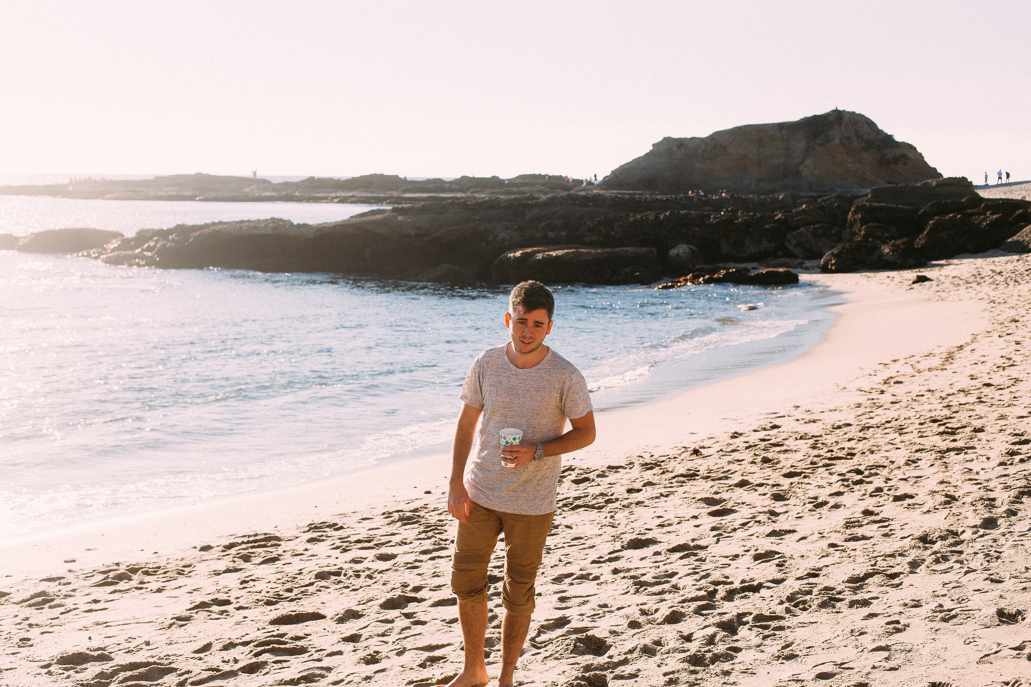 Young man holding a paper cup walking along a sandy beach with rocky outcrops and ocean water.
