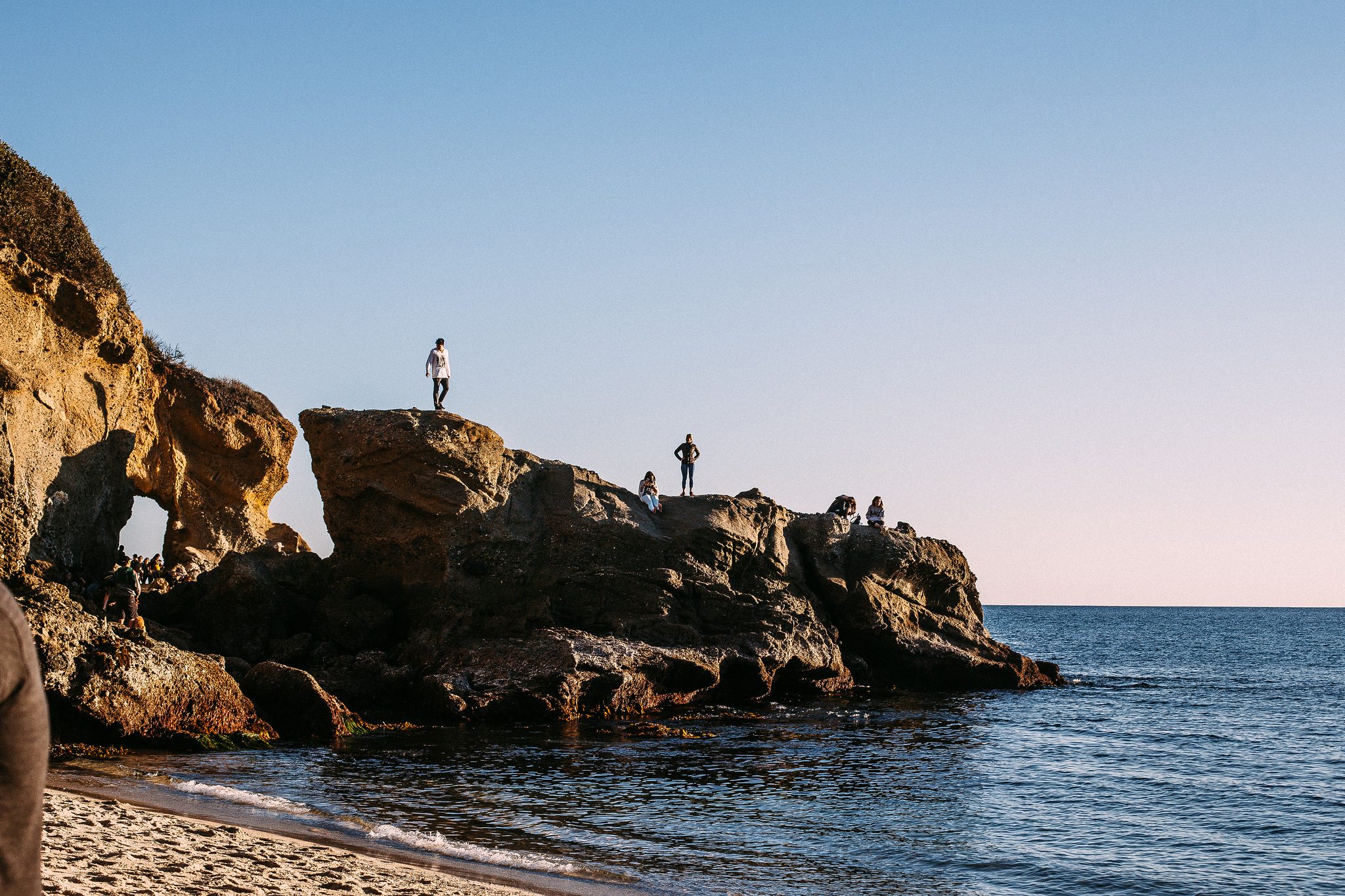 People climbing a rocky sea arch promontory jutting into deep blue ocean water along a sandy beach.