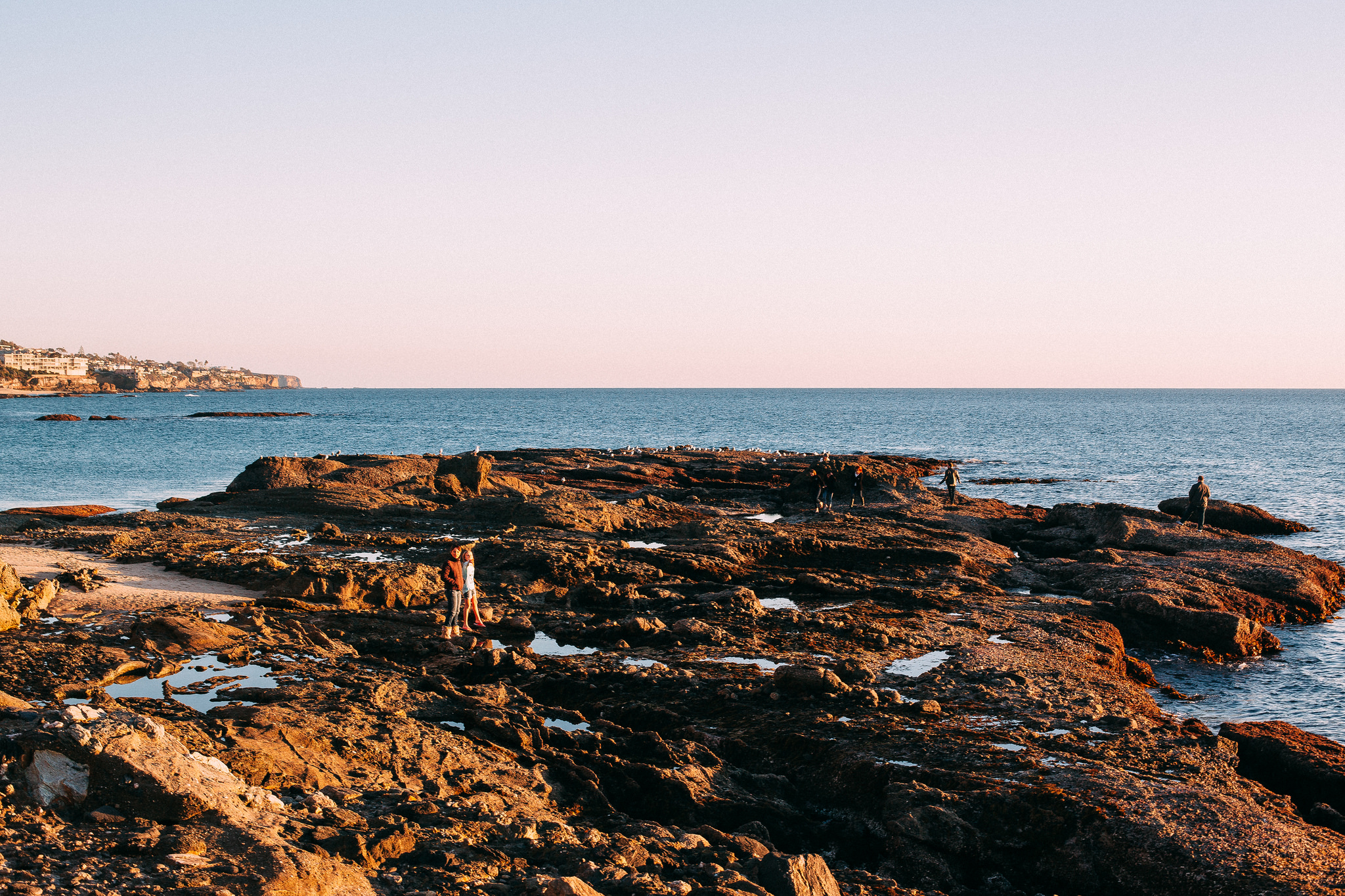 Couples standing on a rocky coastal plateau jutting into calm ocean waters in warm evening sunset light.