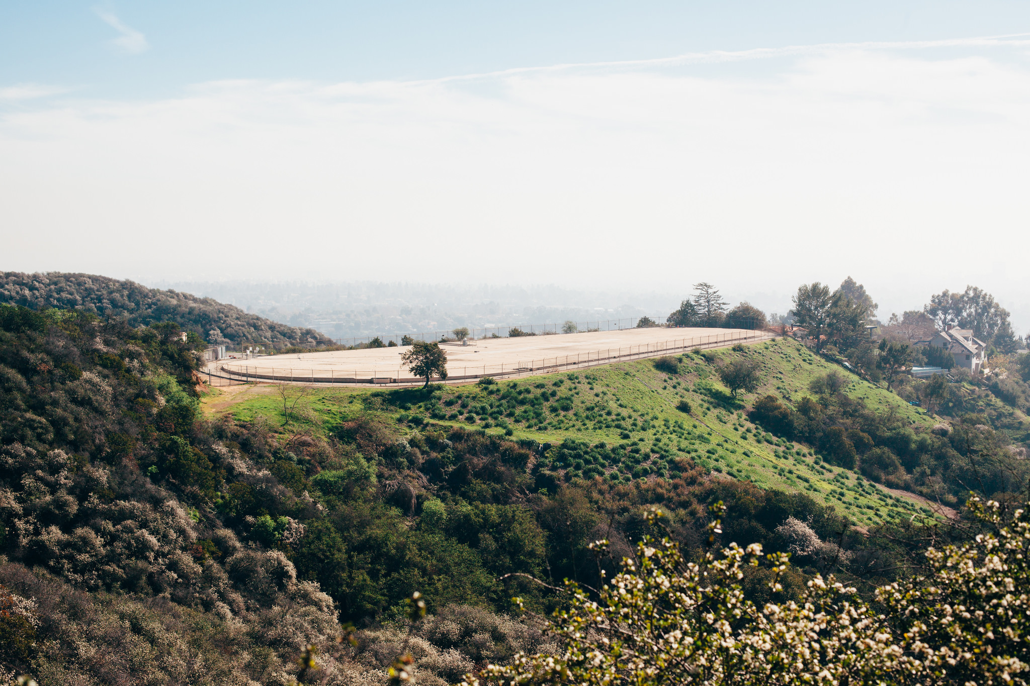 Oval landing pad inside a chain-link fence on a green hillside overlooking a distant city skyline.