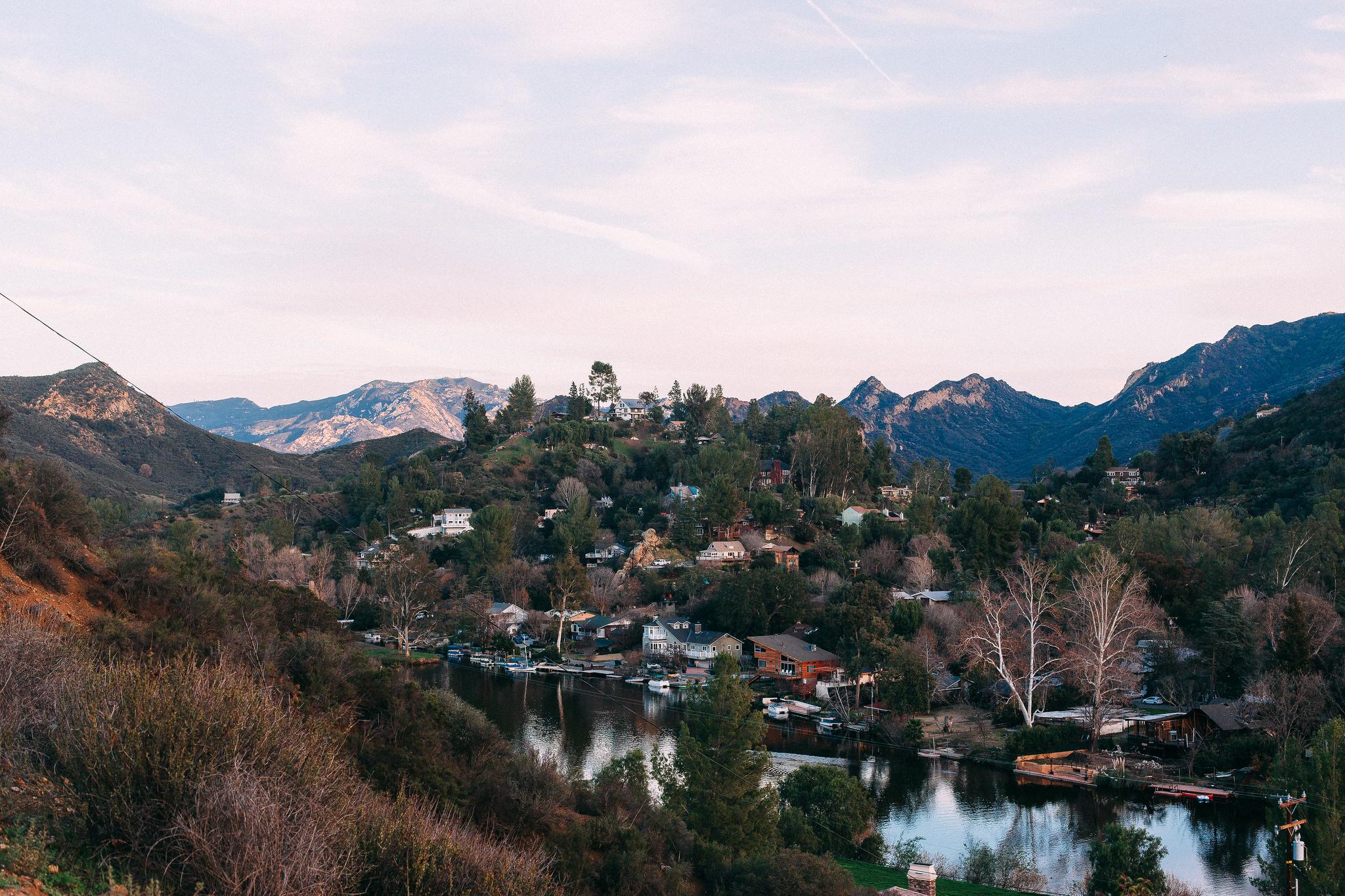 Reflective lake bordered by trees and waterfront homes in rolling hills with distant mountain peaks.
