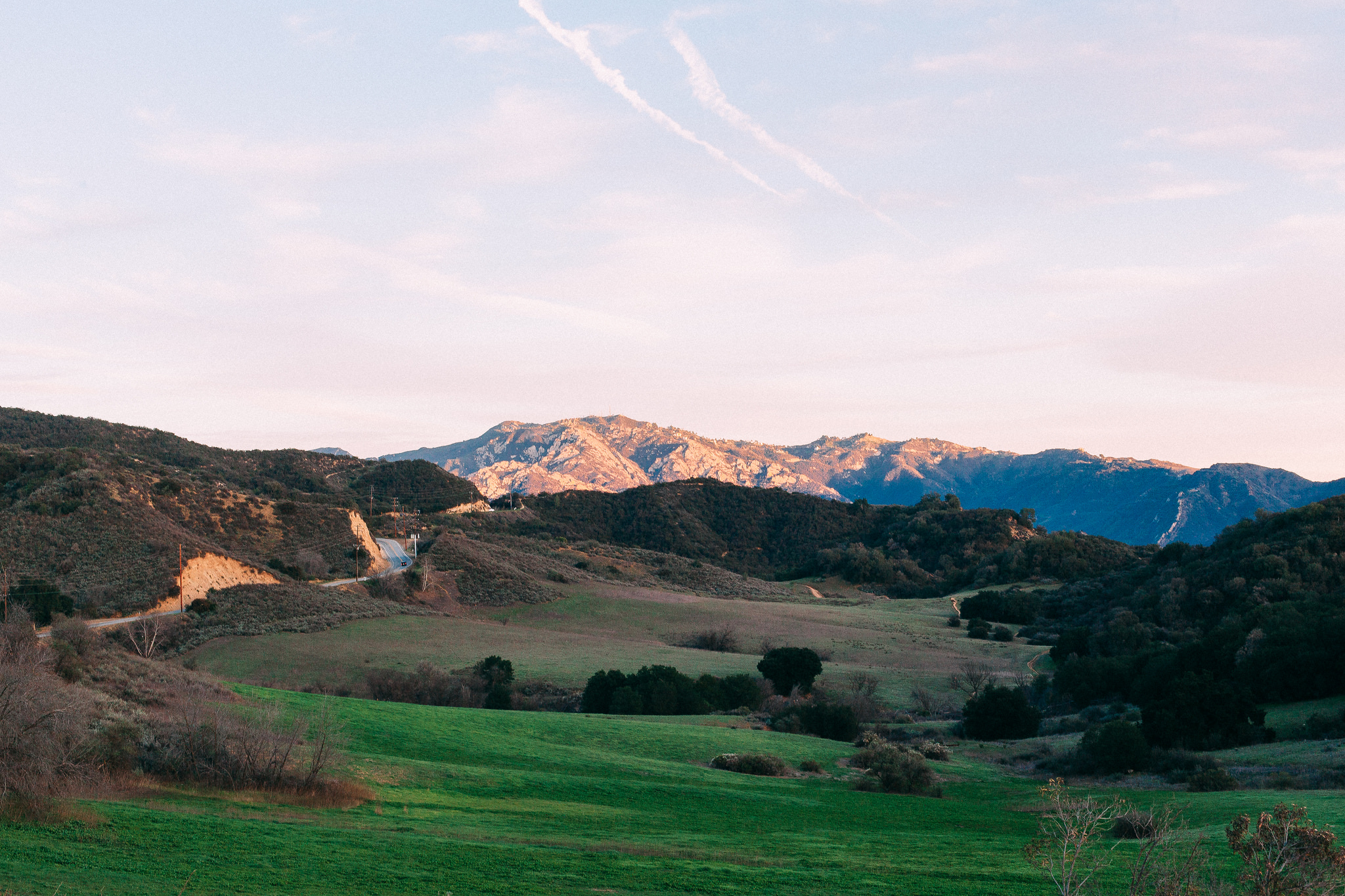 Rolling green hills and a winding country road leading toward sunlit mountain peaks under an evening sky.