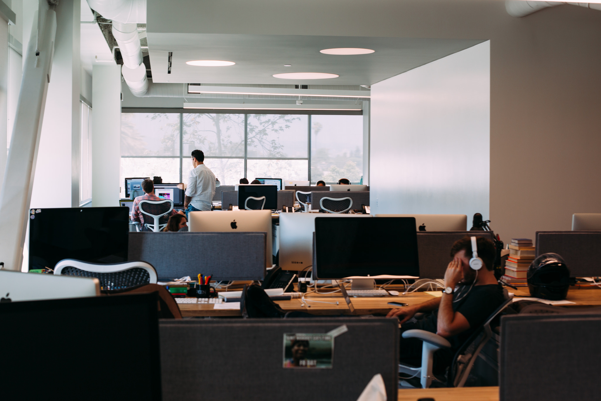Employee wearing white headphones resting his head at a desk while colleagues work in an open office.