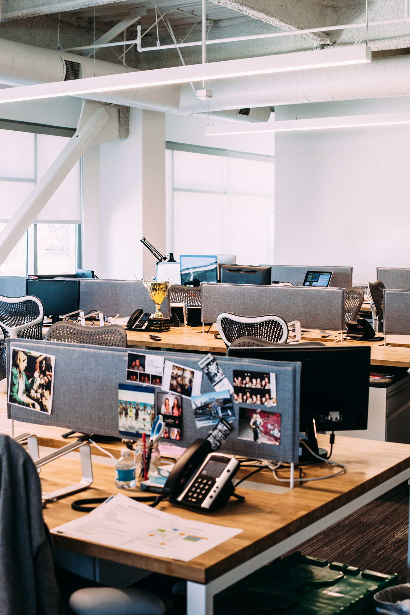 Open office cubicles with wood desks, grey partitions decorated with snapshots, phones, and a trophy.