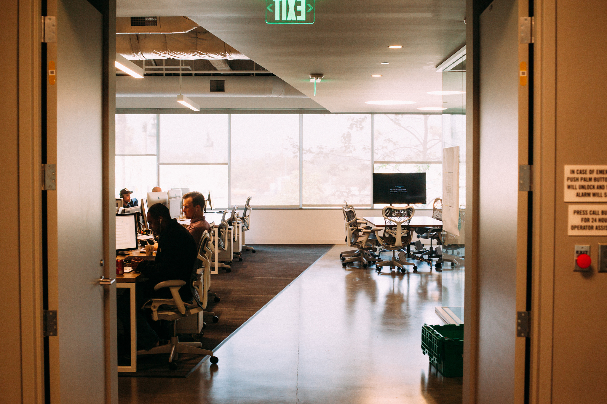 Open doorway view into a modern office with employees at desks, a green exit sign, and meeting room.