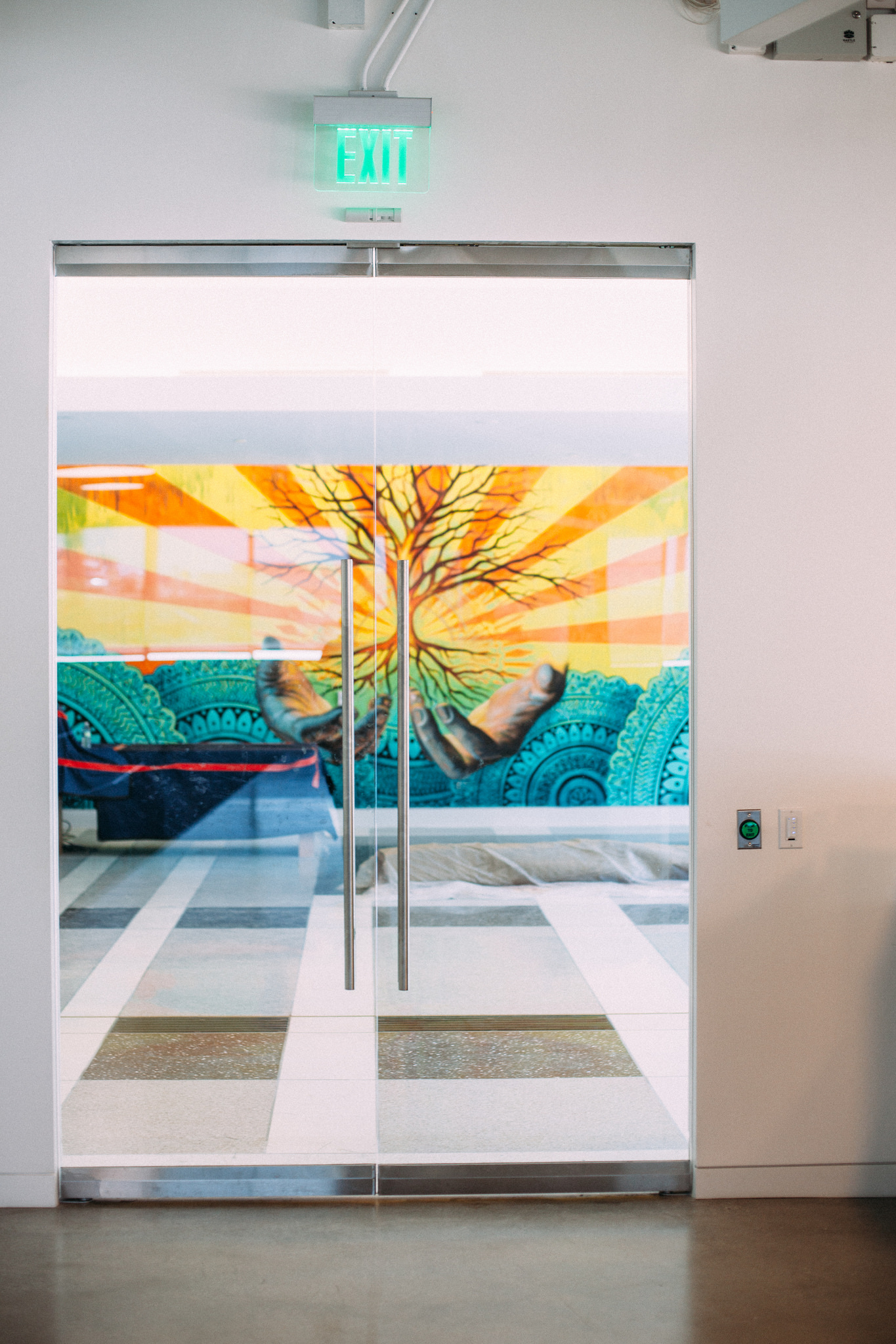 Glass entryway doors beneath an exit sign looking out at a mural of hands holding a tree in sun rays.