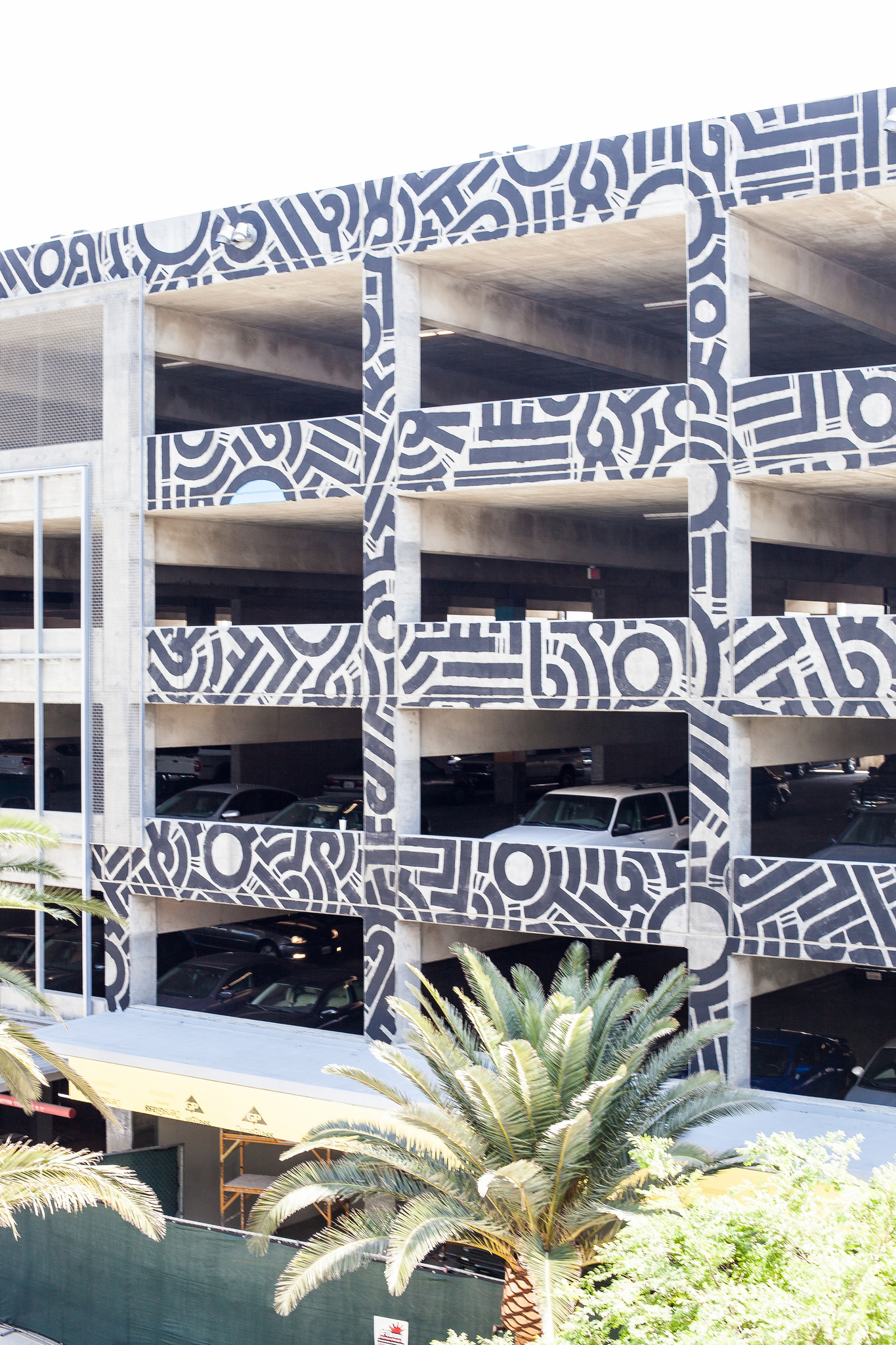 Concrete parking structure painted with bold black-and-white geometric patterns, framed by a palm tree.