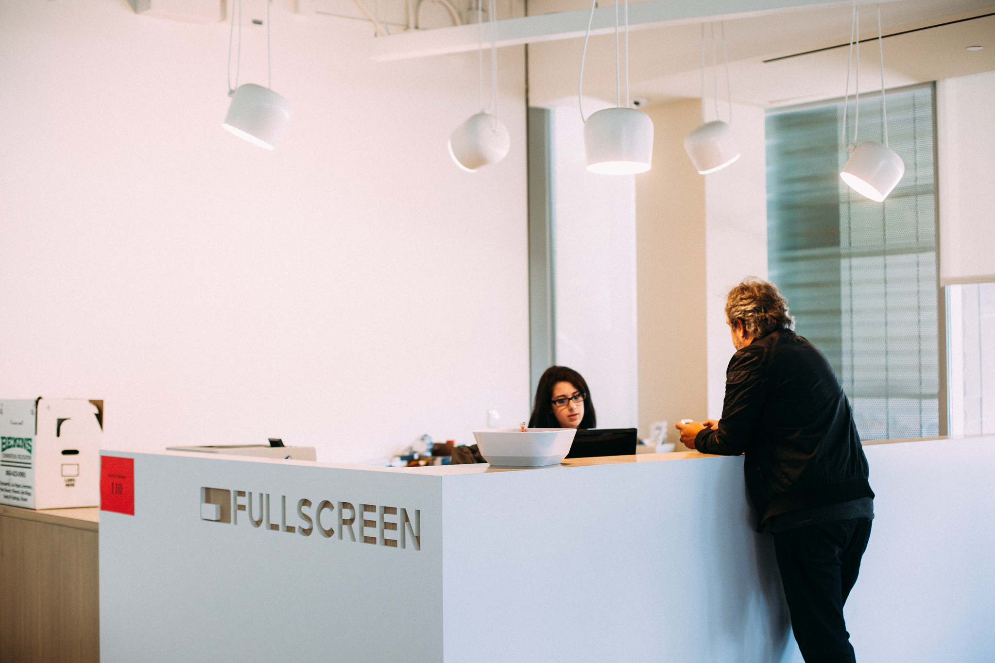 Receptionist behind a white 'FULLSCREEN' desk speaking to a guest in an office lobby with dome lights.