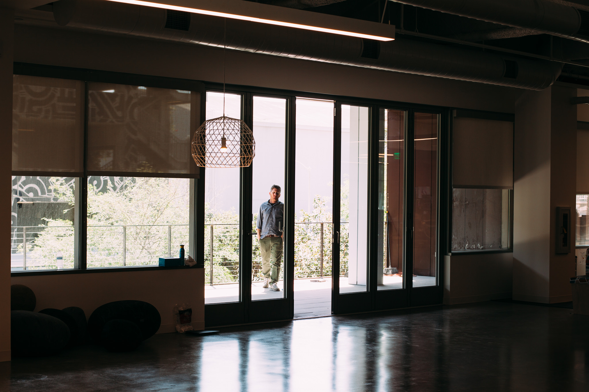 Man on an outdoor office balcony looking into a sunlit open office through glass accordion doors.