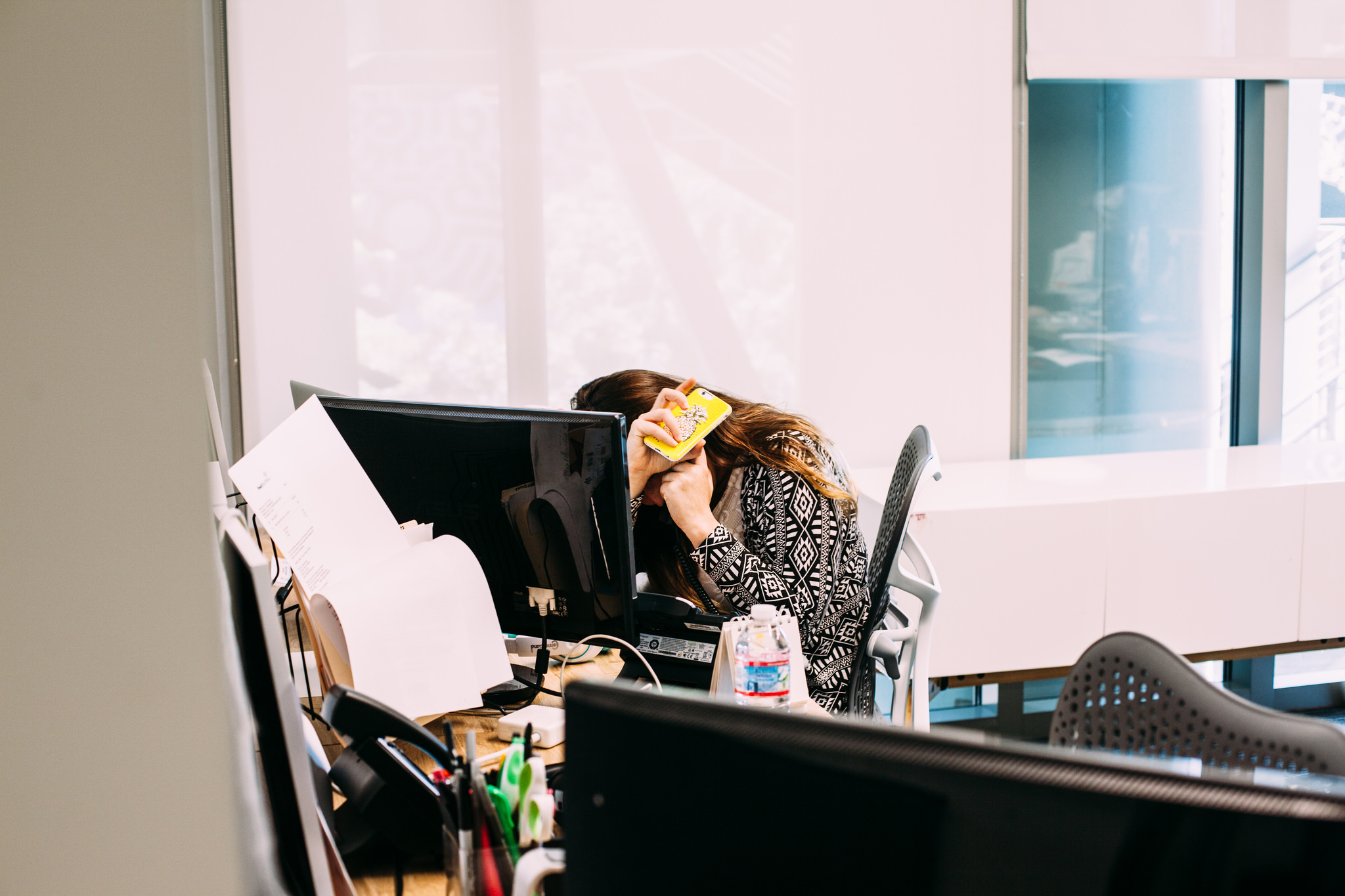 Woman sitting at her office workstation holding a yellow smartphone to her ear with head bowed in frustration.