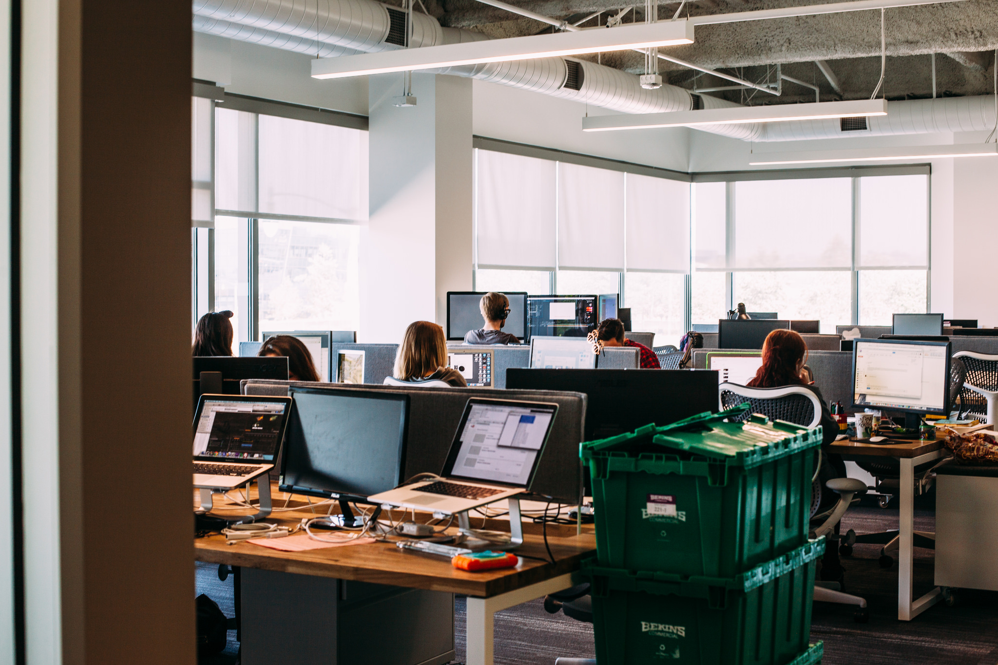 Employees working at dual-monitor workstations in an open office with stacked green crates in foreground.