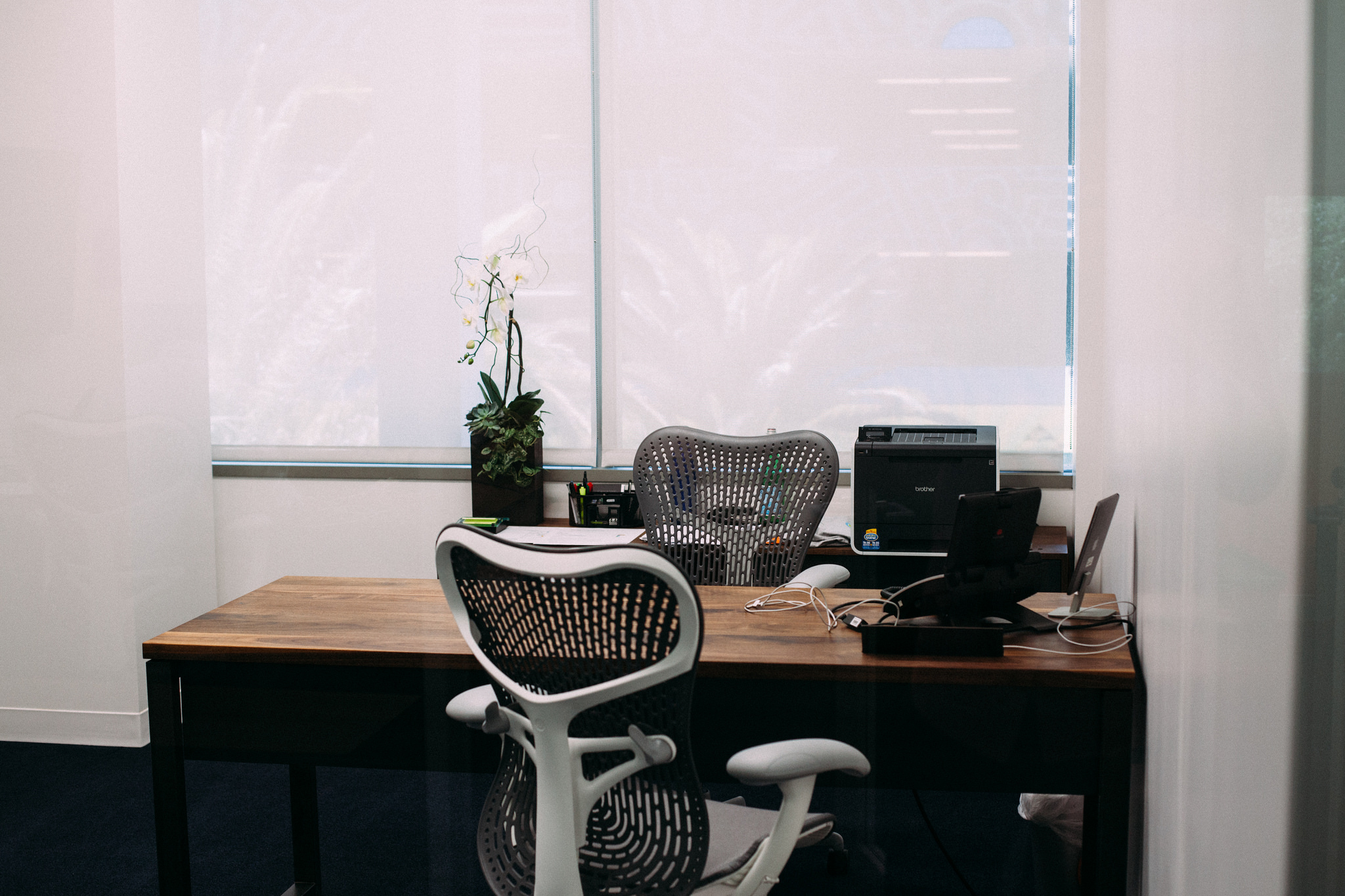 Private office desk with Herman Miller chairs, a potted orchid, printer, and laptop by window blinds.