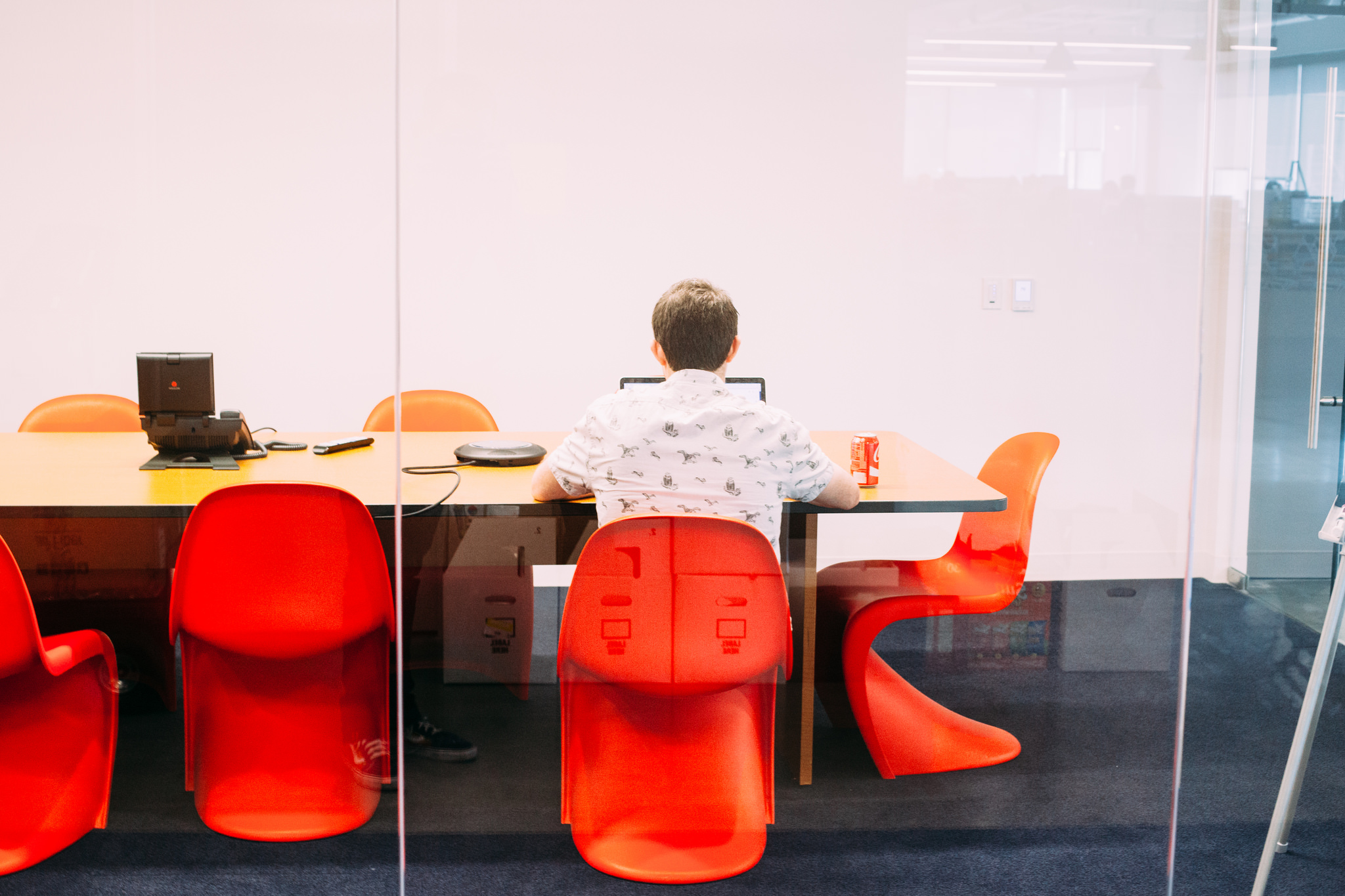 Worker sitting at a long wooden meeting table with a laptop, surrounded by sculptural red chairs.