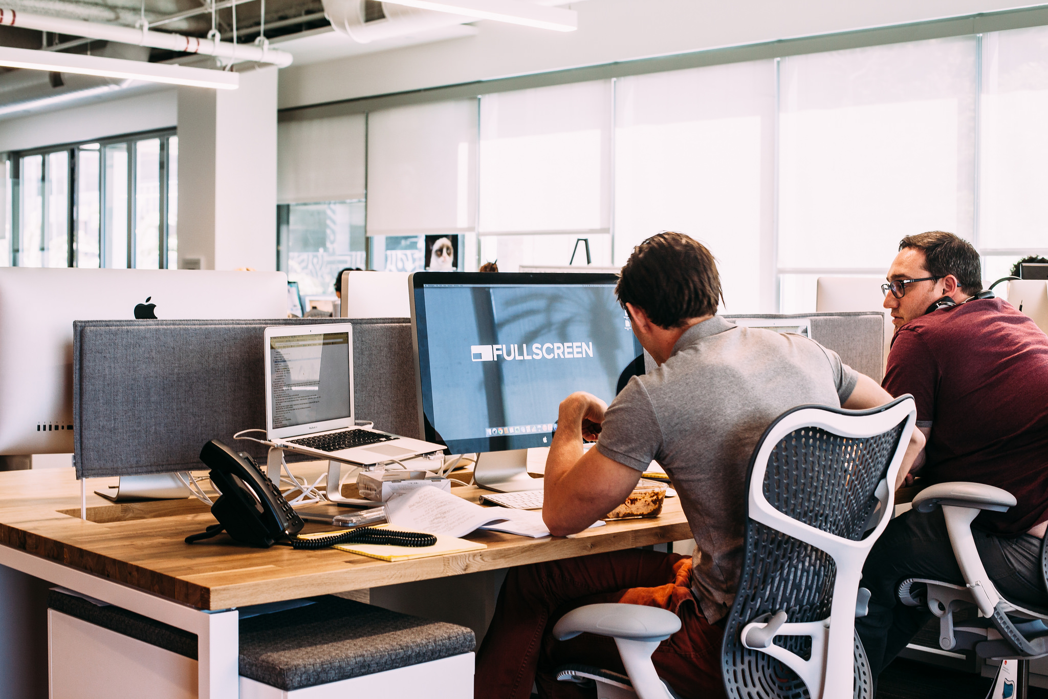 Two coworkers sitting at a wooden desk before a computer monitor displaying the Fullscreen logo.