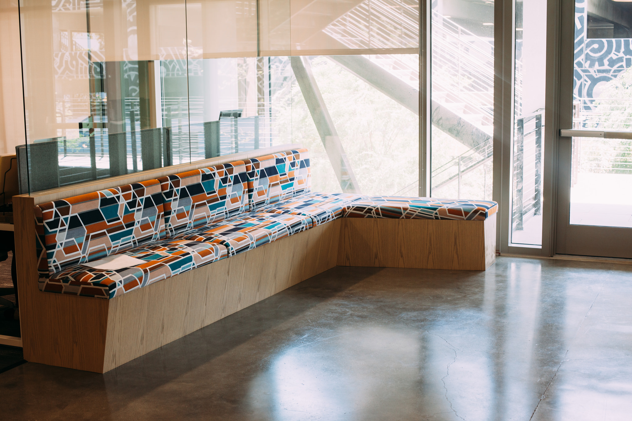 Built-in corner bench with geometric cushions on light wood base beside floor-to-ceiling office windows.