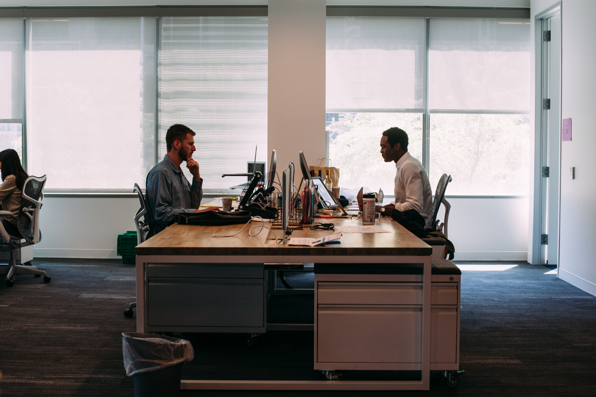 Two coworkers working on laptops opposite each other at a shared wooden desk in an open office.