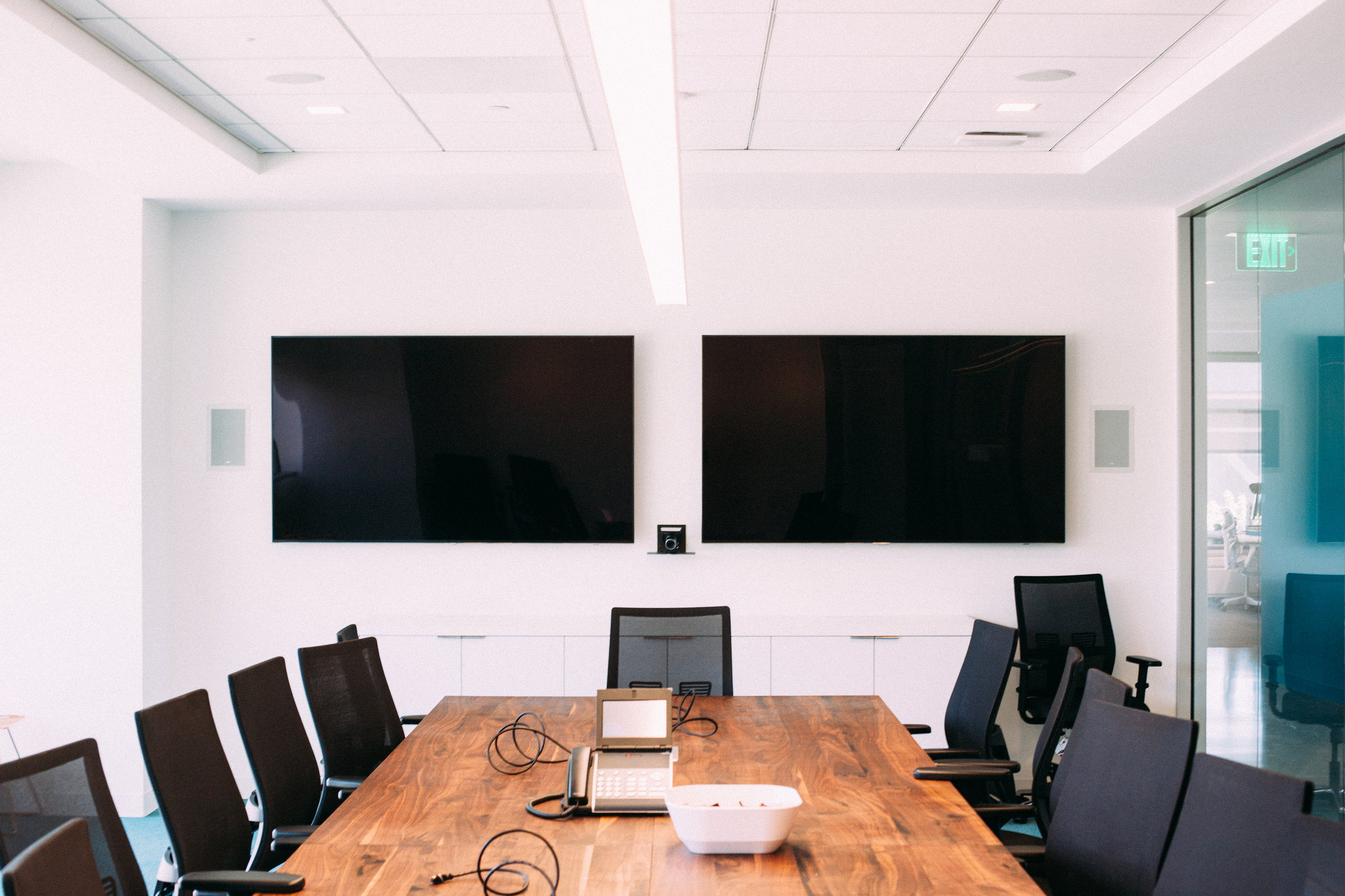 Office boardroom with wall monitors above a long natural edge wood table and black ergonomic chairs.