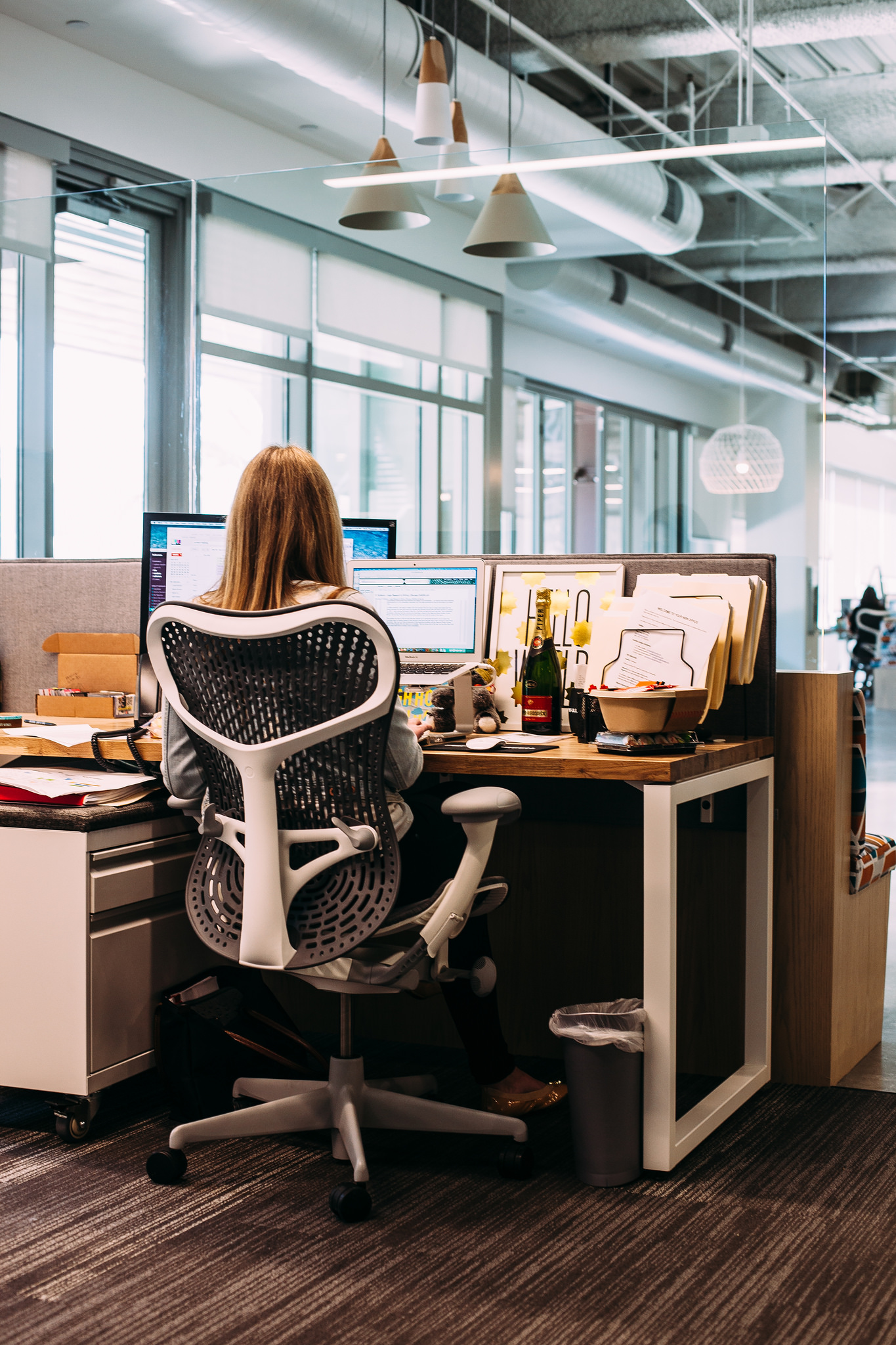 Woman sitting at an office desk working on dual monitors in an ergonomic white desk chair.