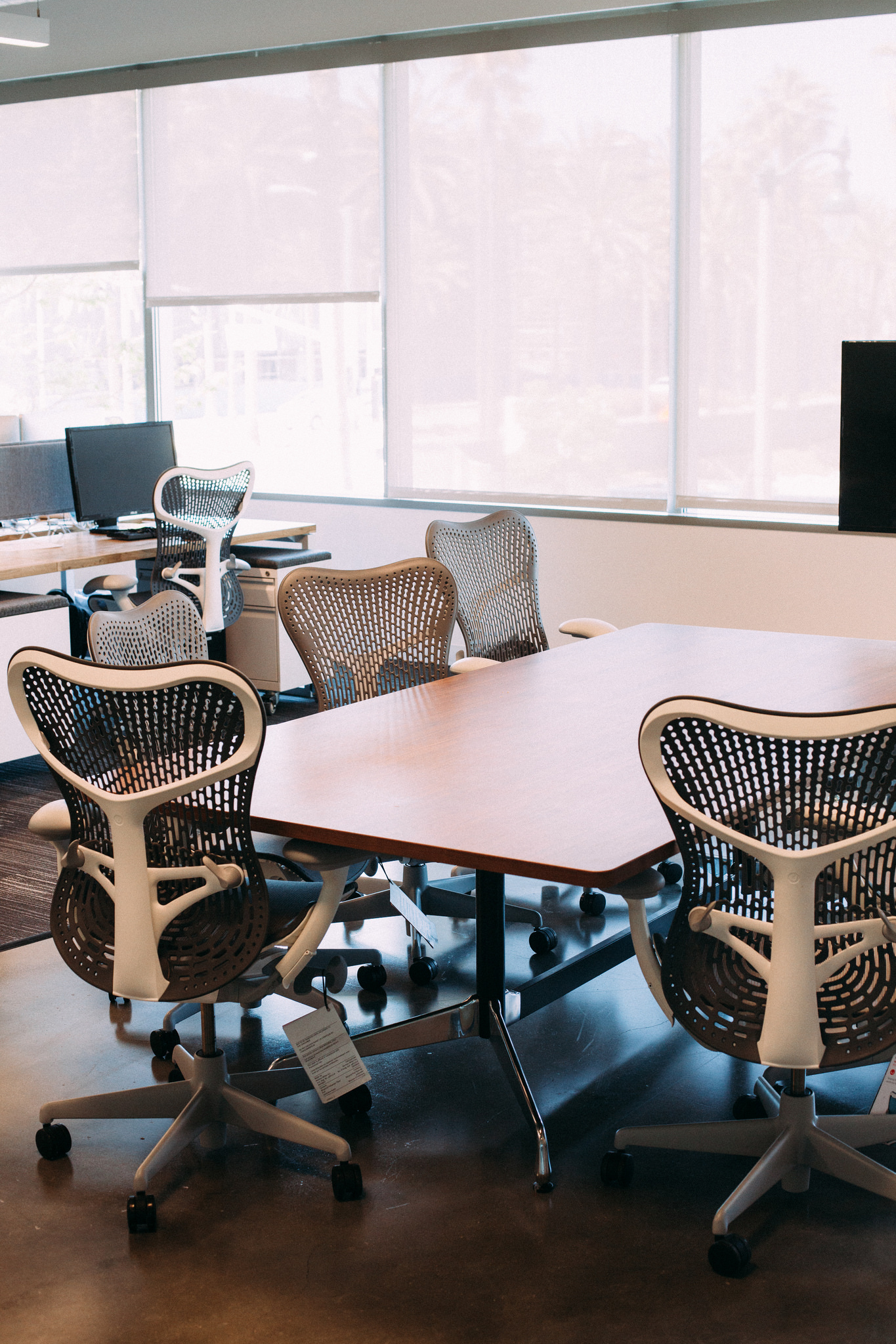 Conference room with light wood table surrounded by grey ergonomic chairs near sunlit windows with shades.