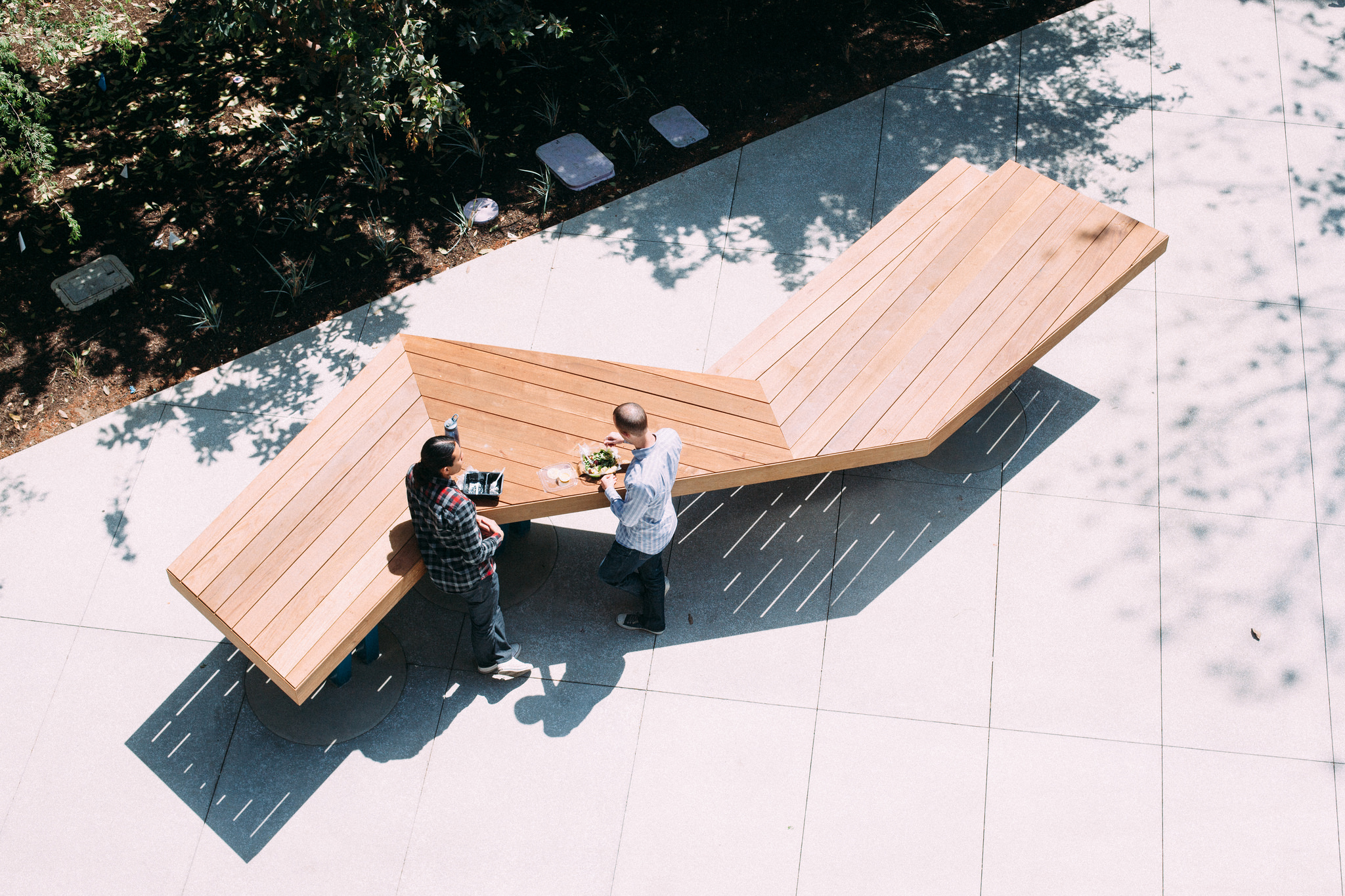 High-angle view of two people eating lunch at an angular wooden outdoor bench on a paved patio.