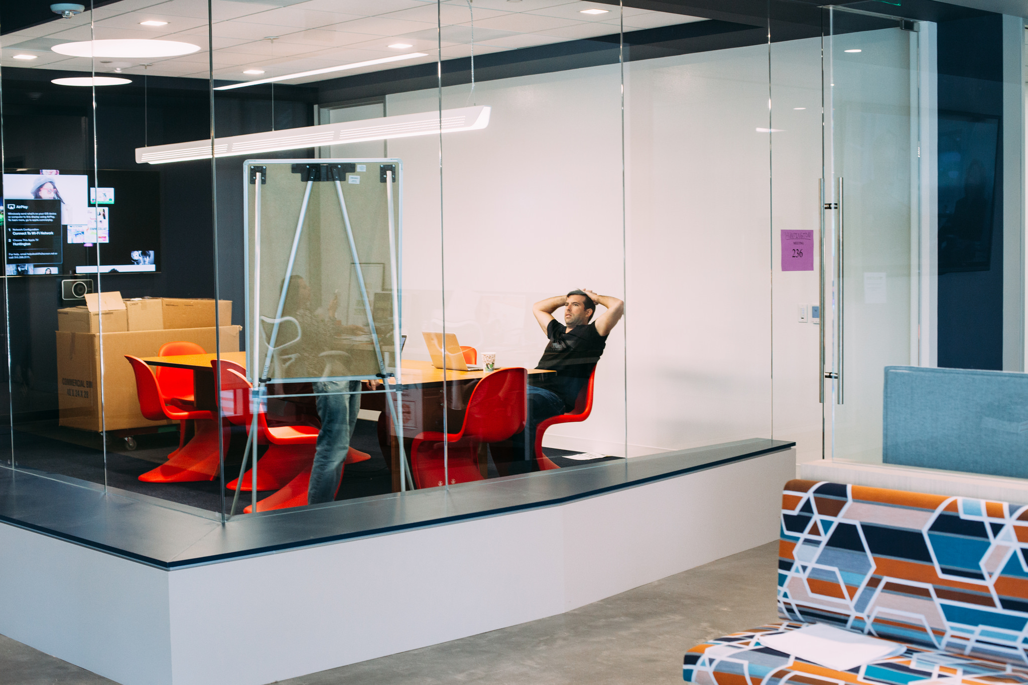 Man in a black shirt leans back in a red chair at a meeting table inside a glass conference room.