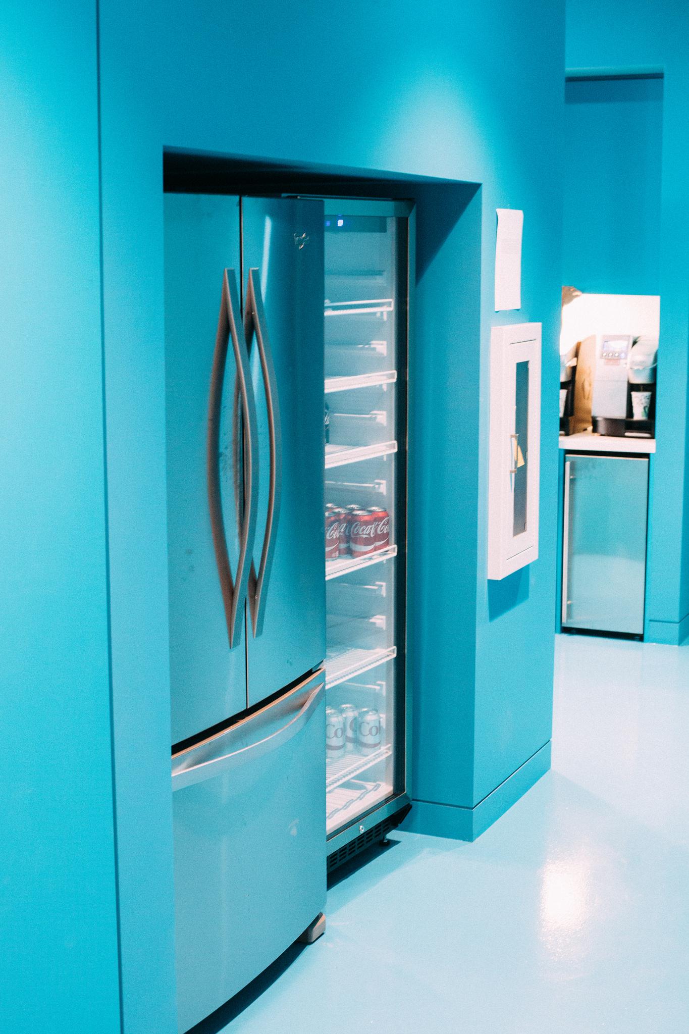 Glass-door refrigerator showing Coca-Cola cans set into vibrant turquoise office kitchenette walls.