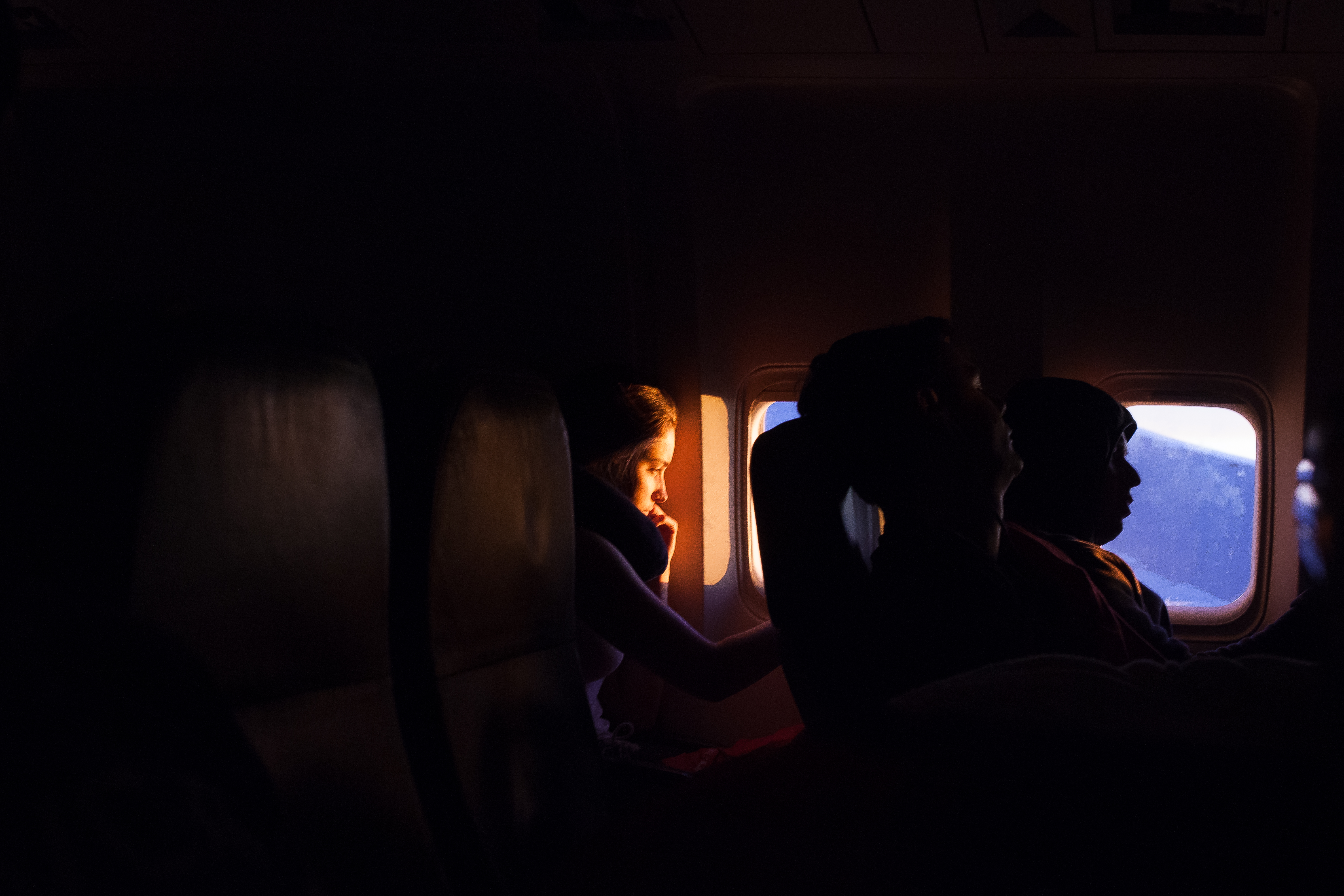 Shaft of warm golden sunlight illuminating a young woman's face inside a dark airplane cabin during flight, shot with a Canon 5D Mark II.