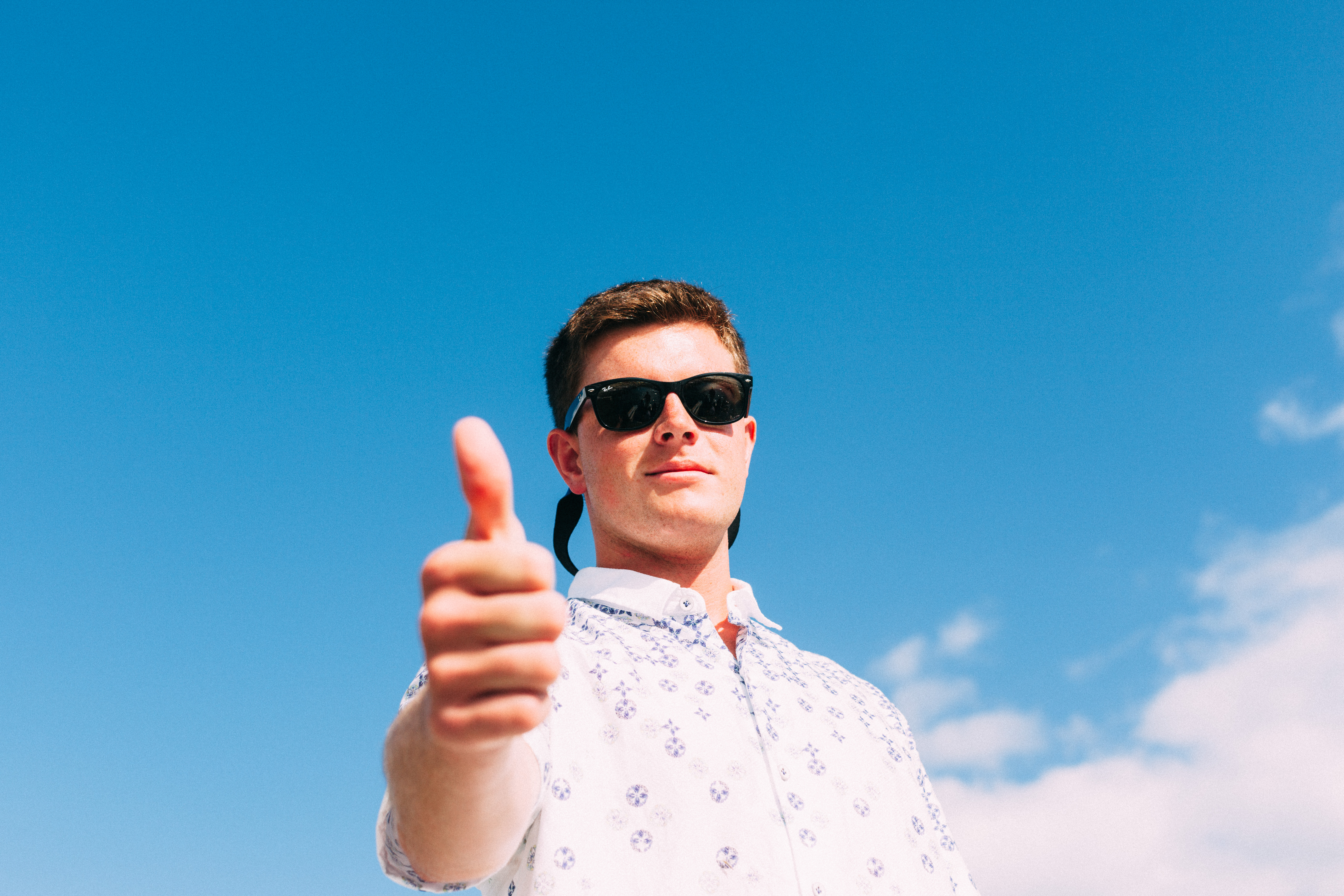 Young man wearing sunglasses and a white printed button-down shirt giving a confident thumbs-up against a vivid clear blue sky, shot with a Canon 5D Mark II.