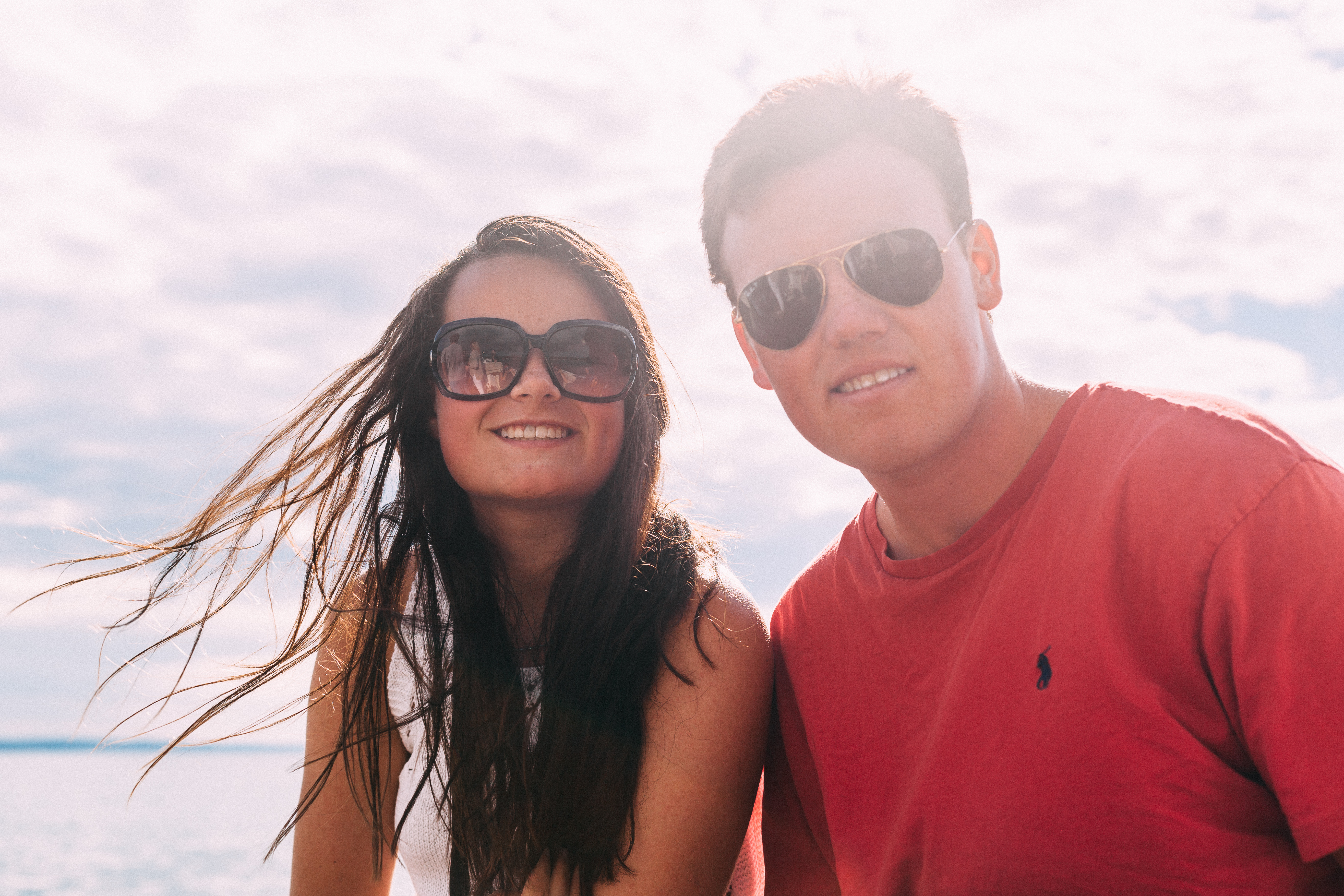 Smiling young woman with windblown brown hair in dark sunglasses leaning close to a smiling young man in aviators and a red tee on a boat, shot with a Canon 5D Mark II.