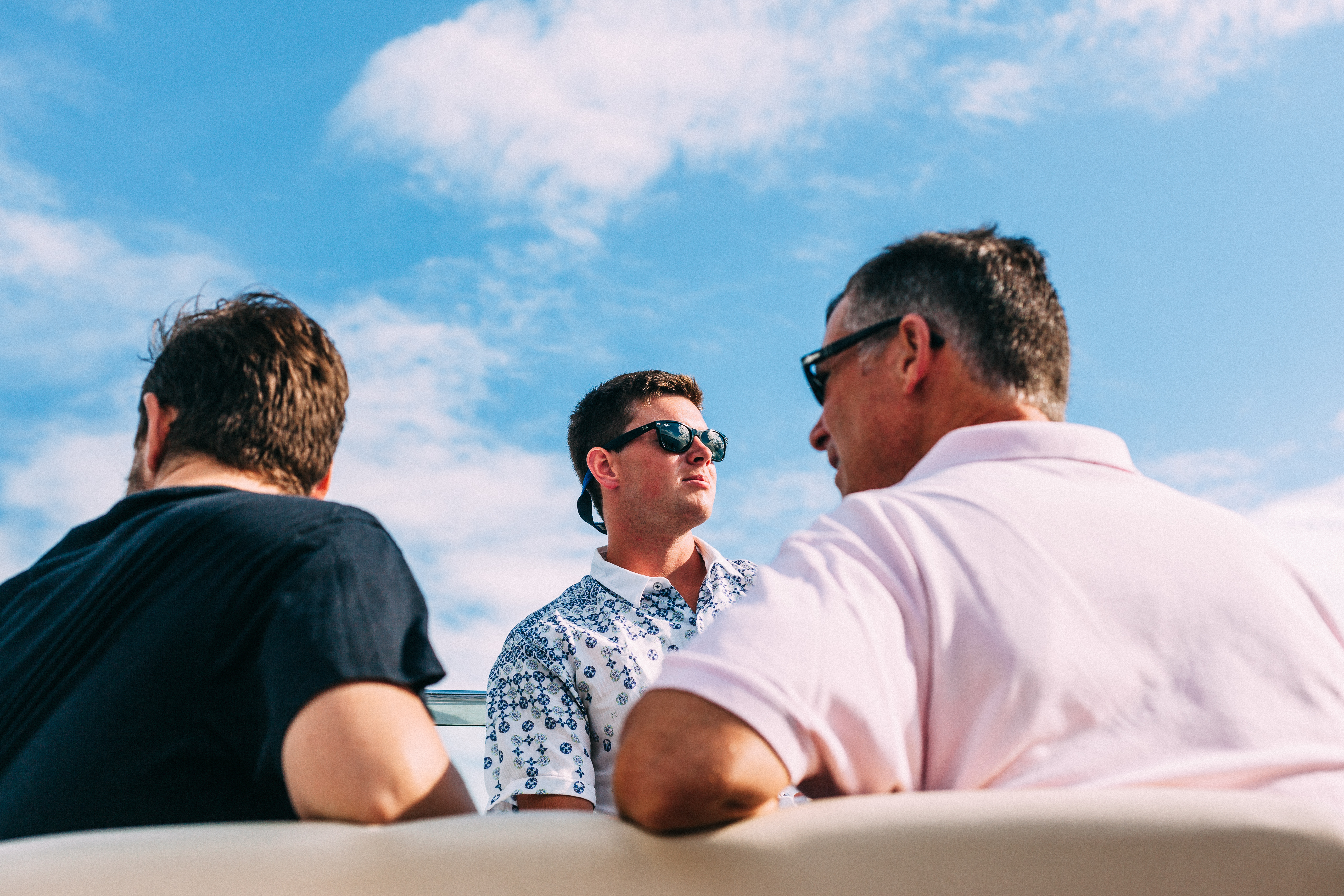 Low-angle candid of three friends seated together on a boat under a bright blue sky, with a young man in a patterned shirt looking into the distance, shot with a Canon 5D Mark II.