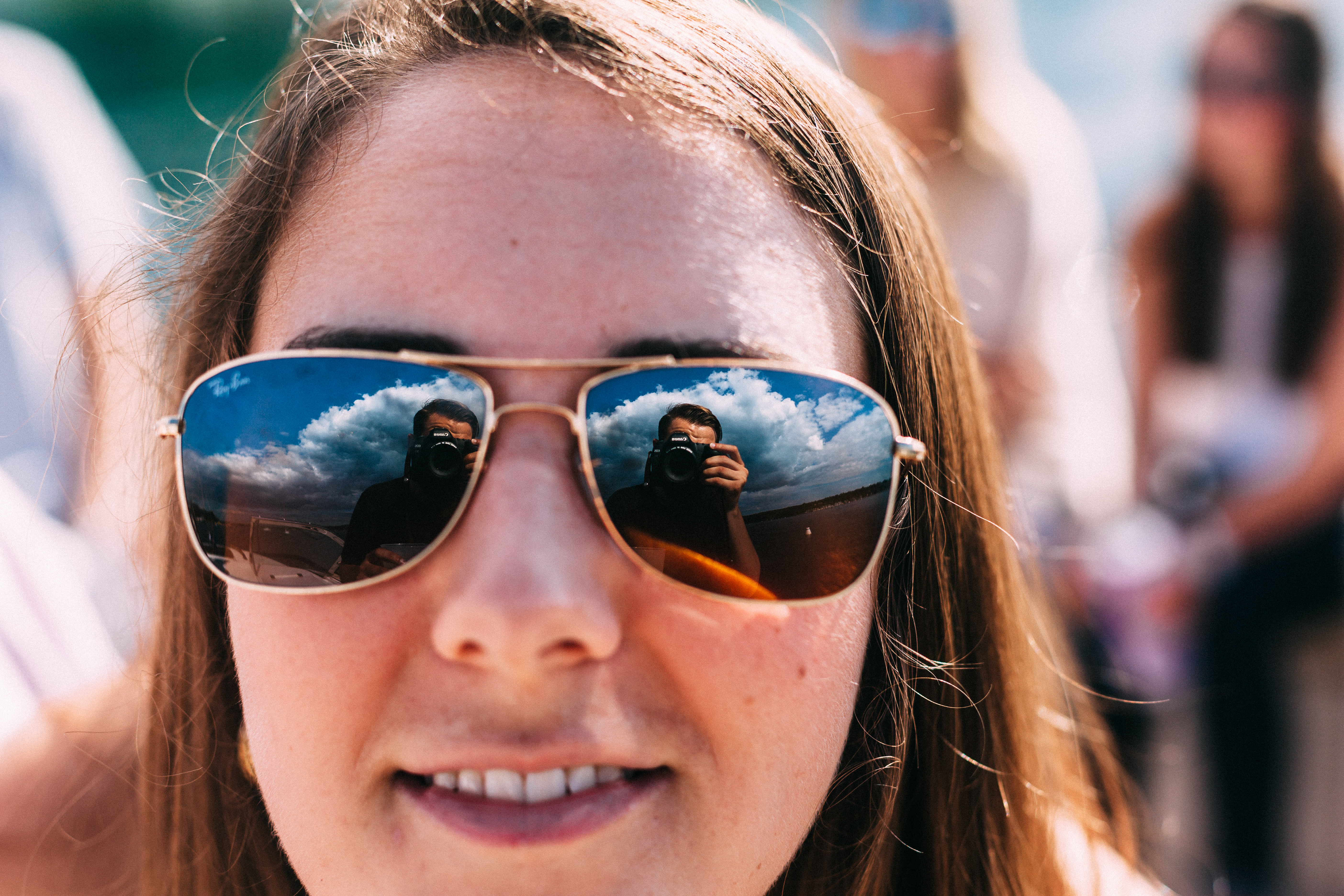 Close-up portrait of a smiling woman in gold aviator sunglasses reflecting the photographer holding a Canon DSLR camera against a blue sky, shot with a Canon 5D Mark II.