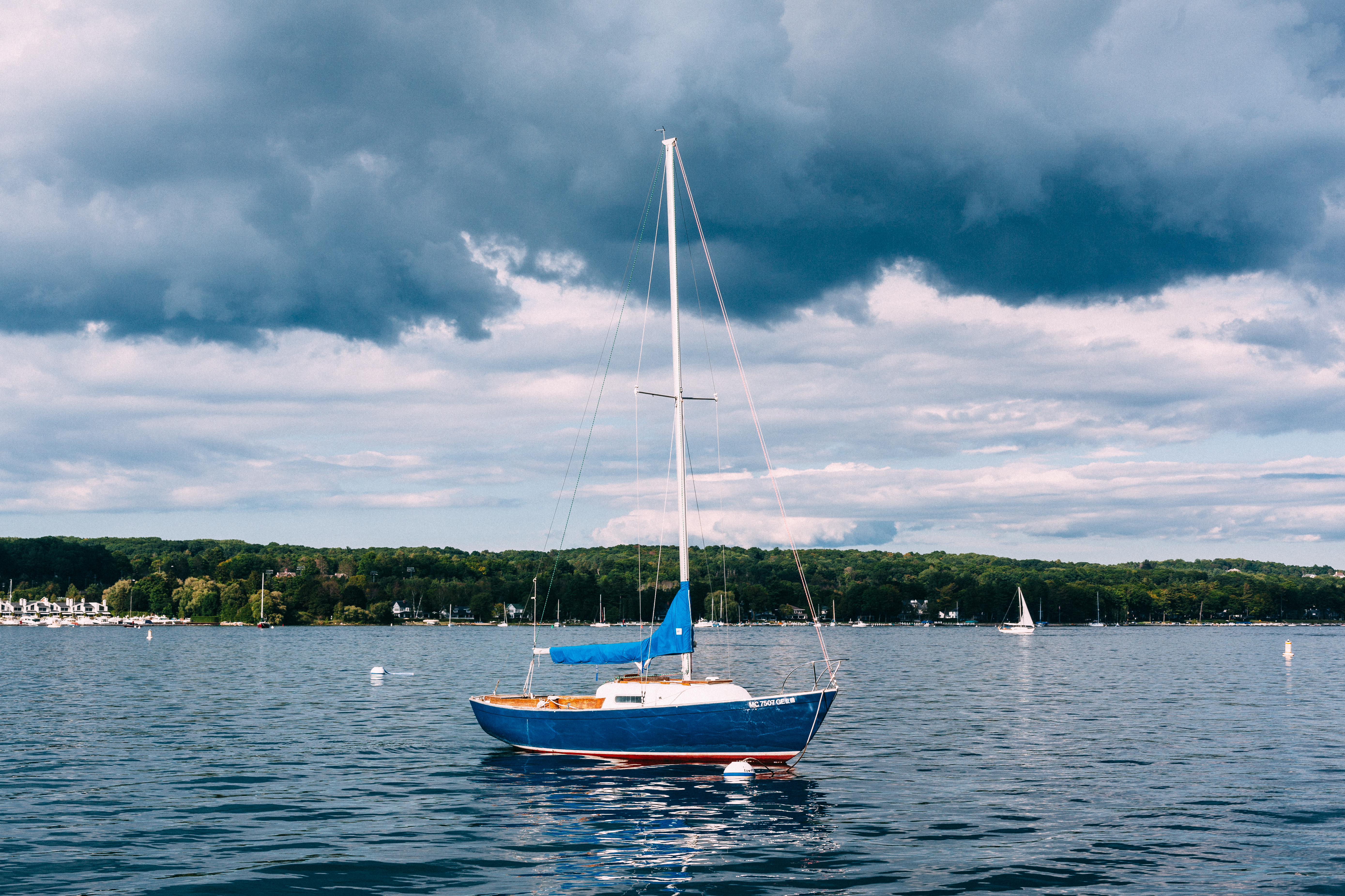 Small blue-hulled sailboat moored on calm lake waters beneath dramatic dark storm clouds and a forested shoreline, shot with a Canon 5D Mark II.