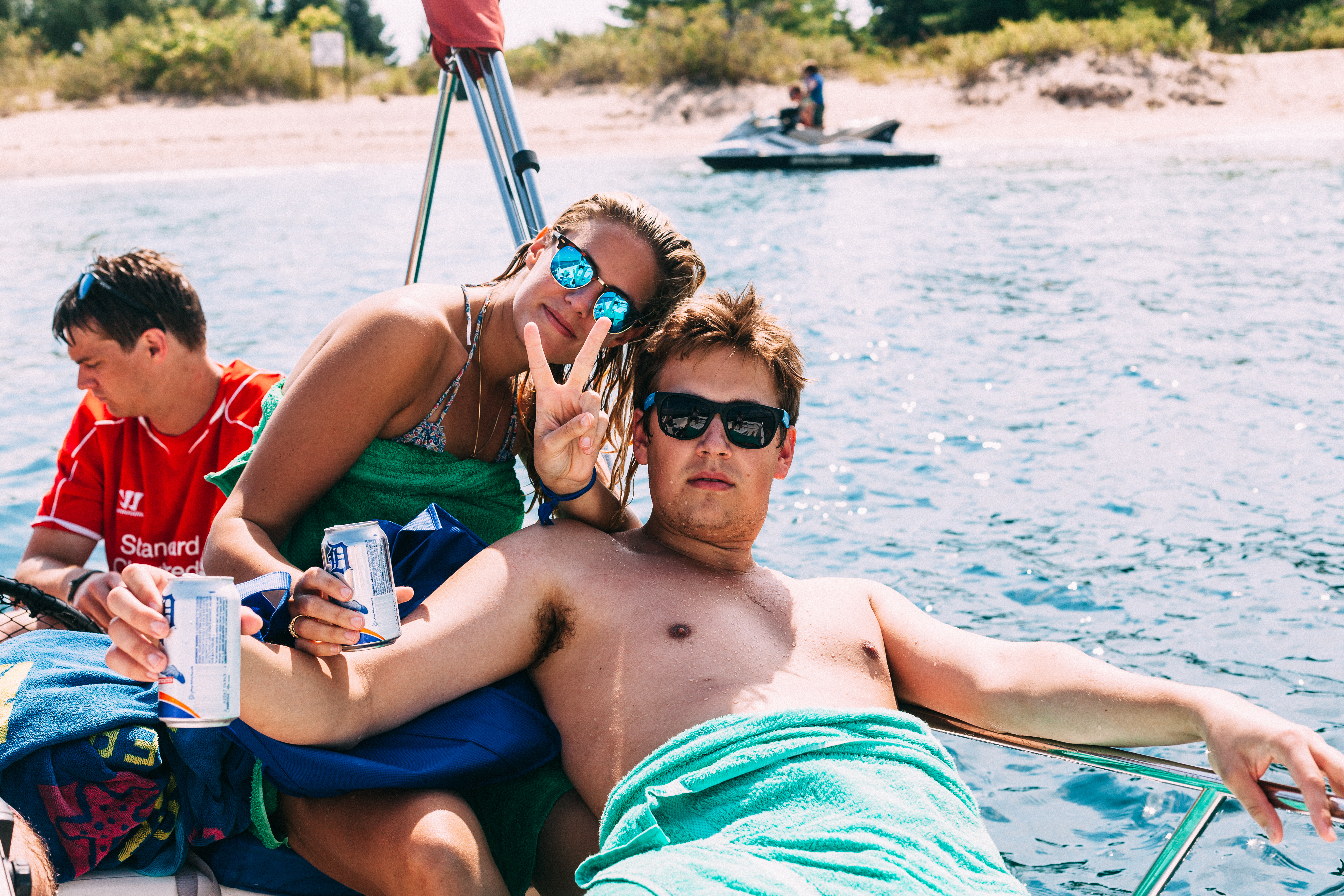 Friends on a lake boat, with a man wrapped in a turquoise towel and woman flashing a peace sign.