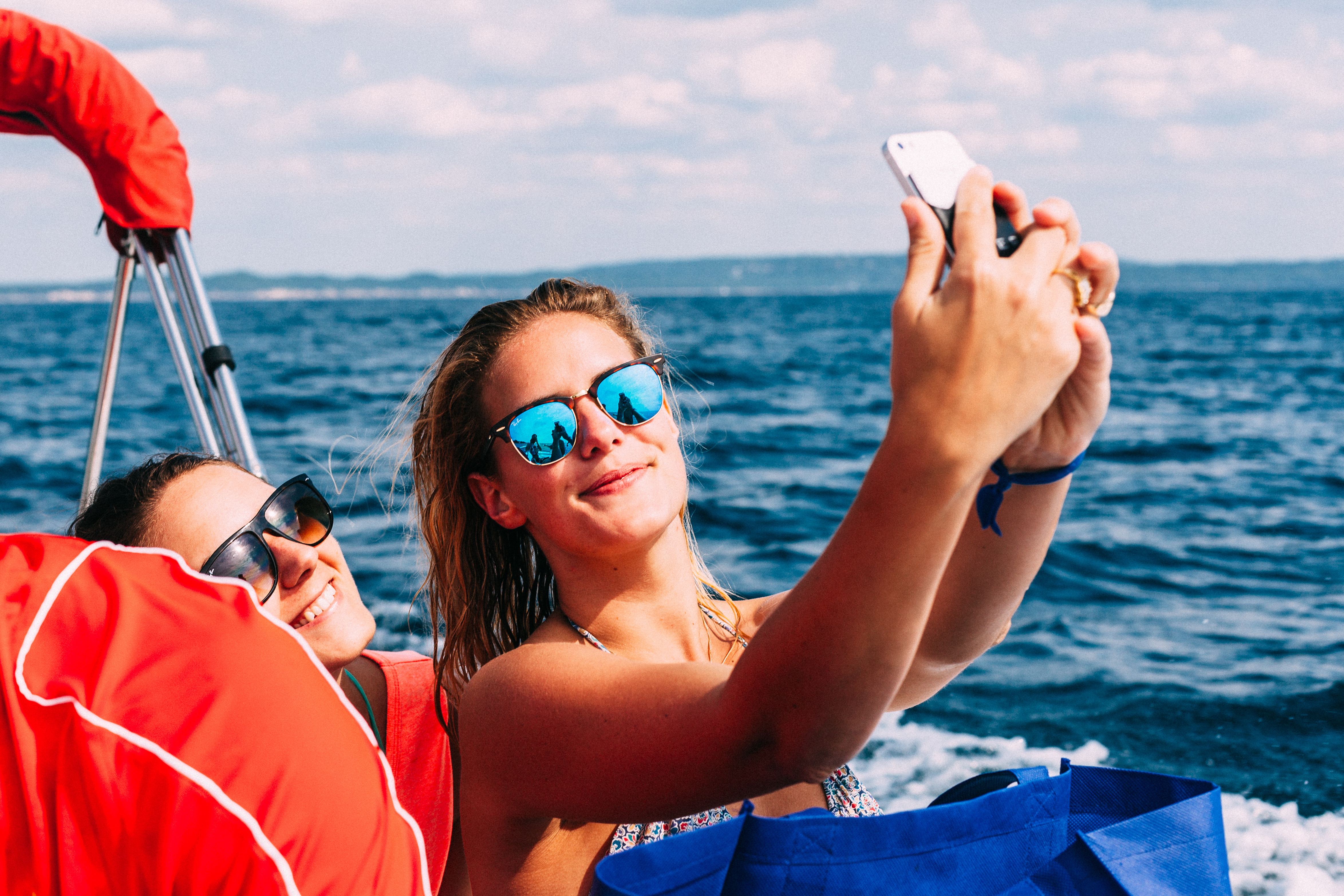 Two smiling women on a moving boat taking a phone selfie against deep blue lake waters.