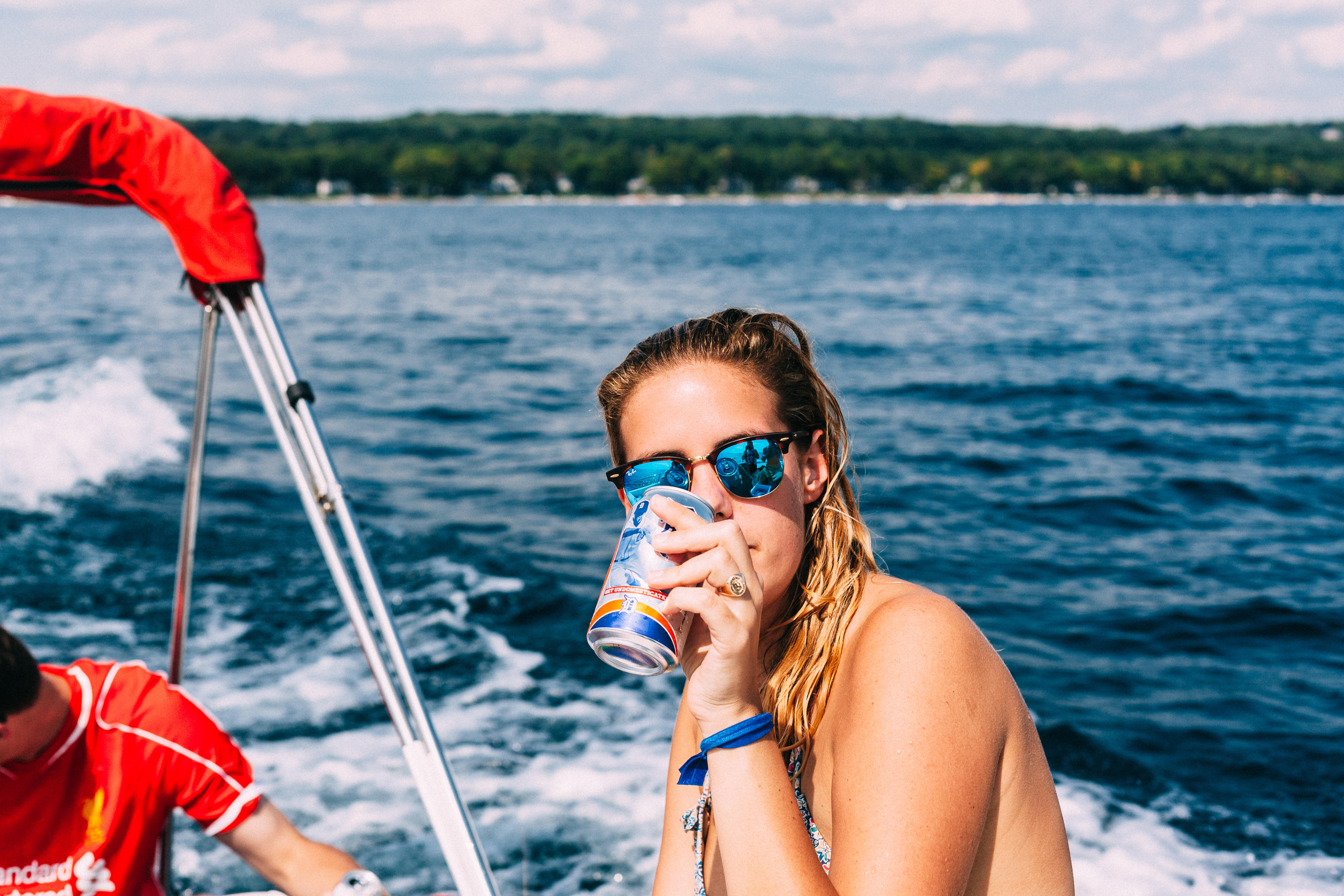 Woman in blue mirrored sunglasses sipping a canned drink on a boat riding across open blue water.