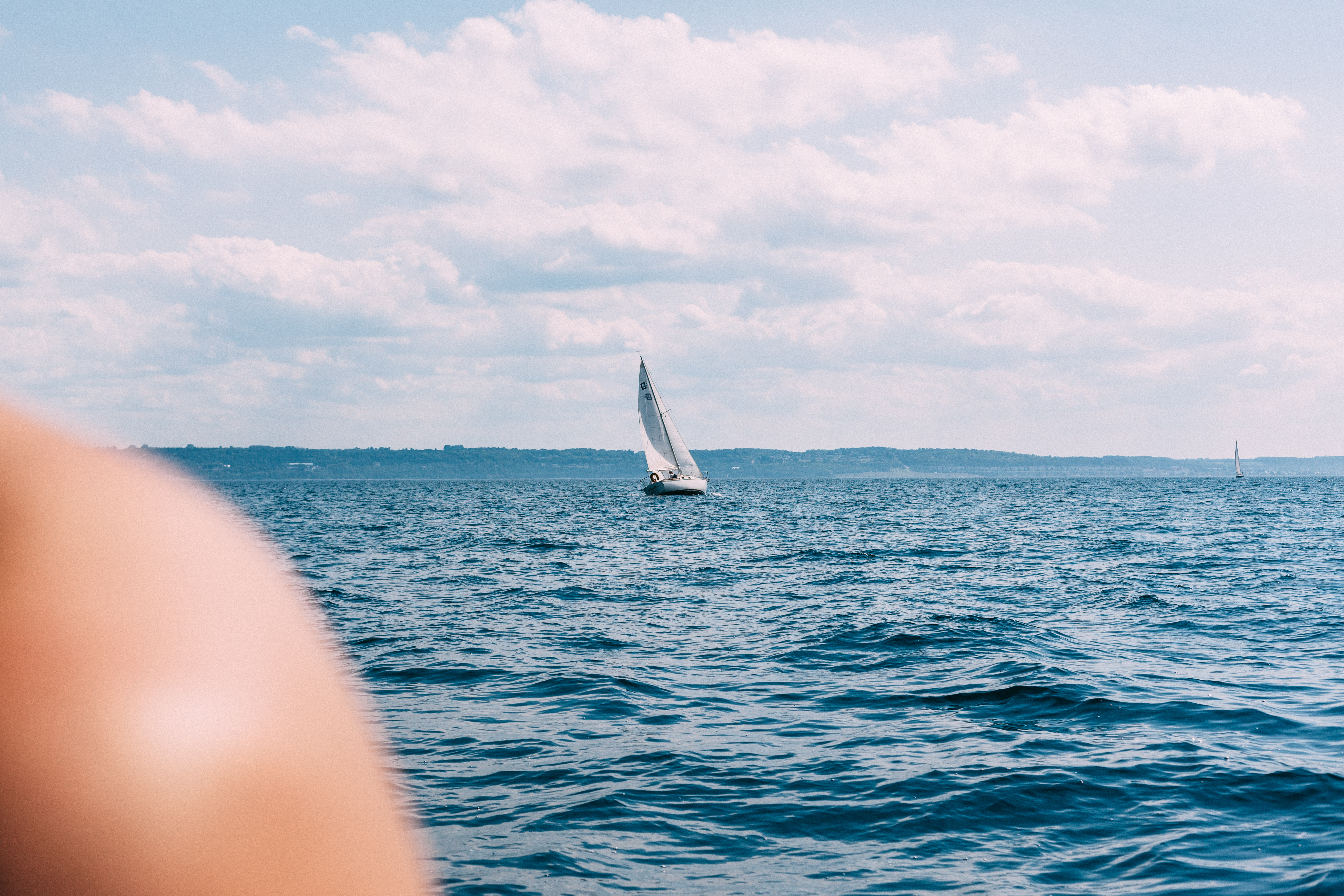 Distant white sailboat sailing across choppy deep blue lake waters under soft blue sky with fluffy white clouds.