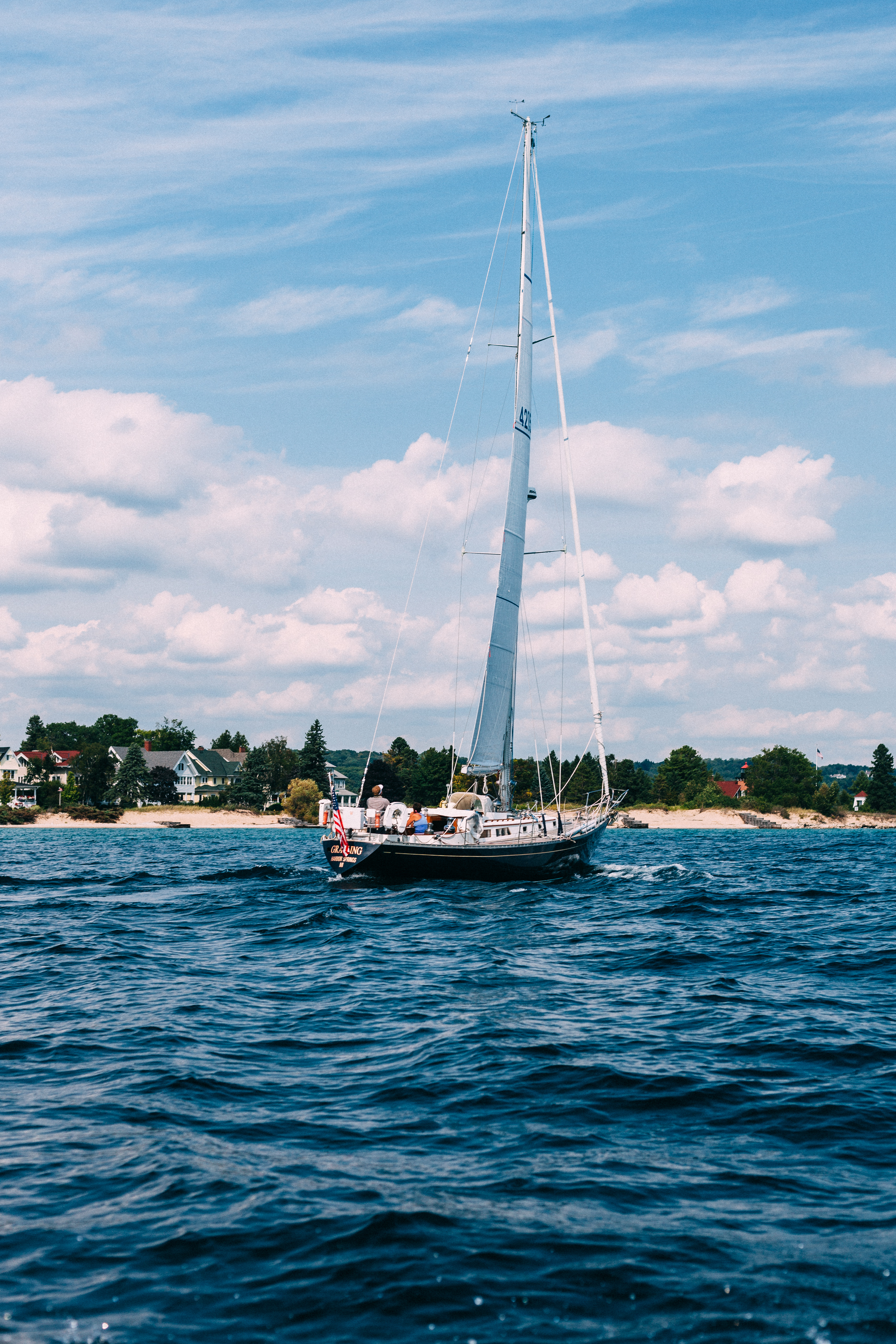Cruising sailboat named Grayling motoring across blue lake water toward a tree-lined shoreline.