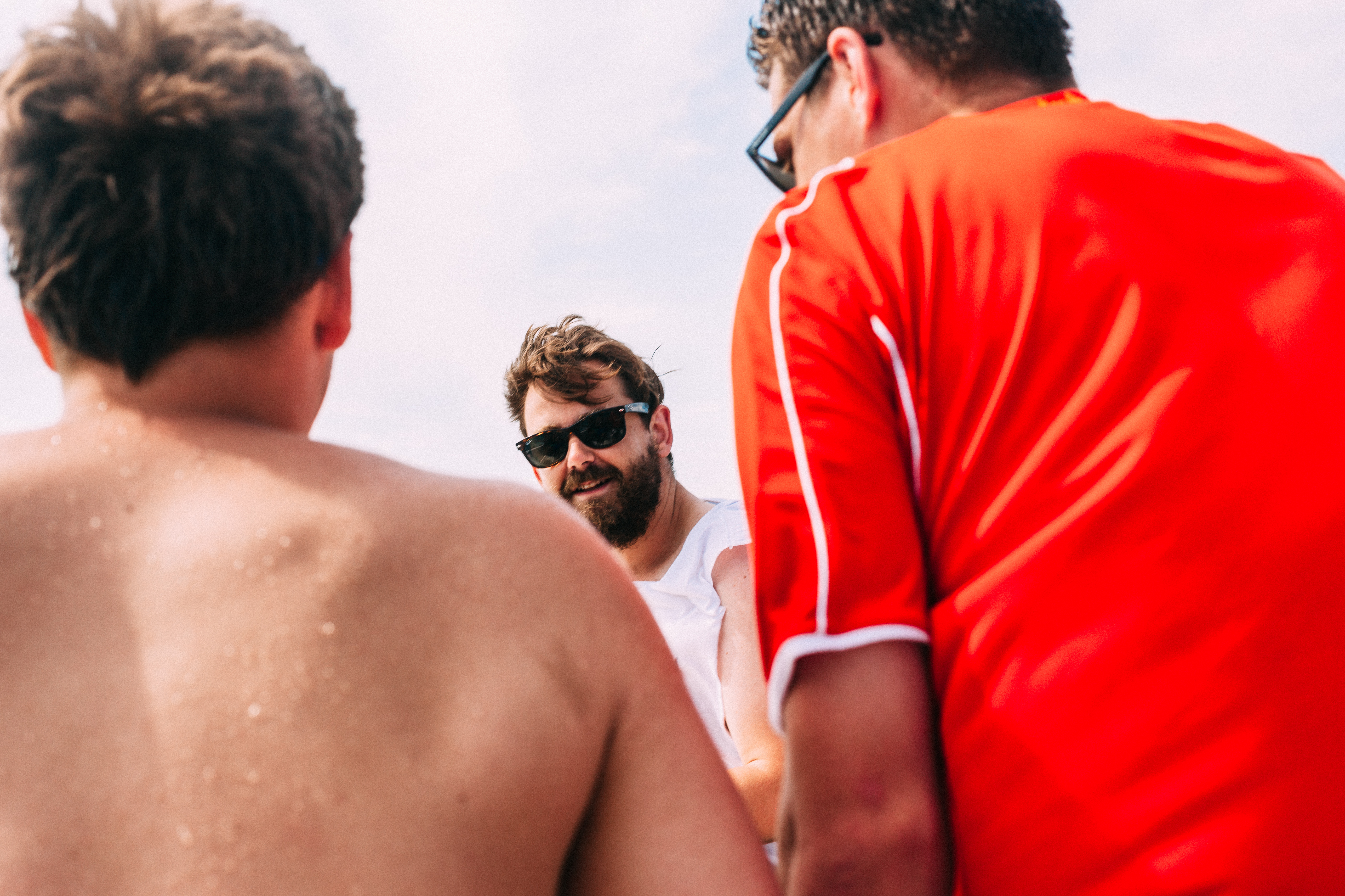 Bearded young man in sunglasses smiling with friends in casual athletic wear on a sunny boat deck.