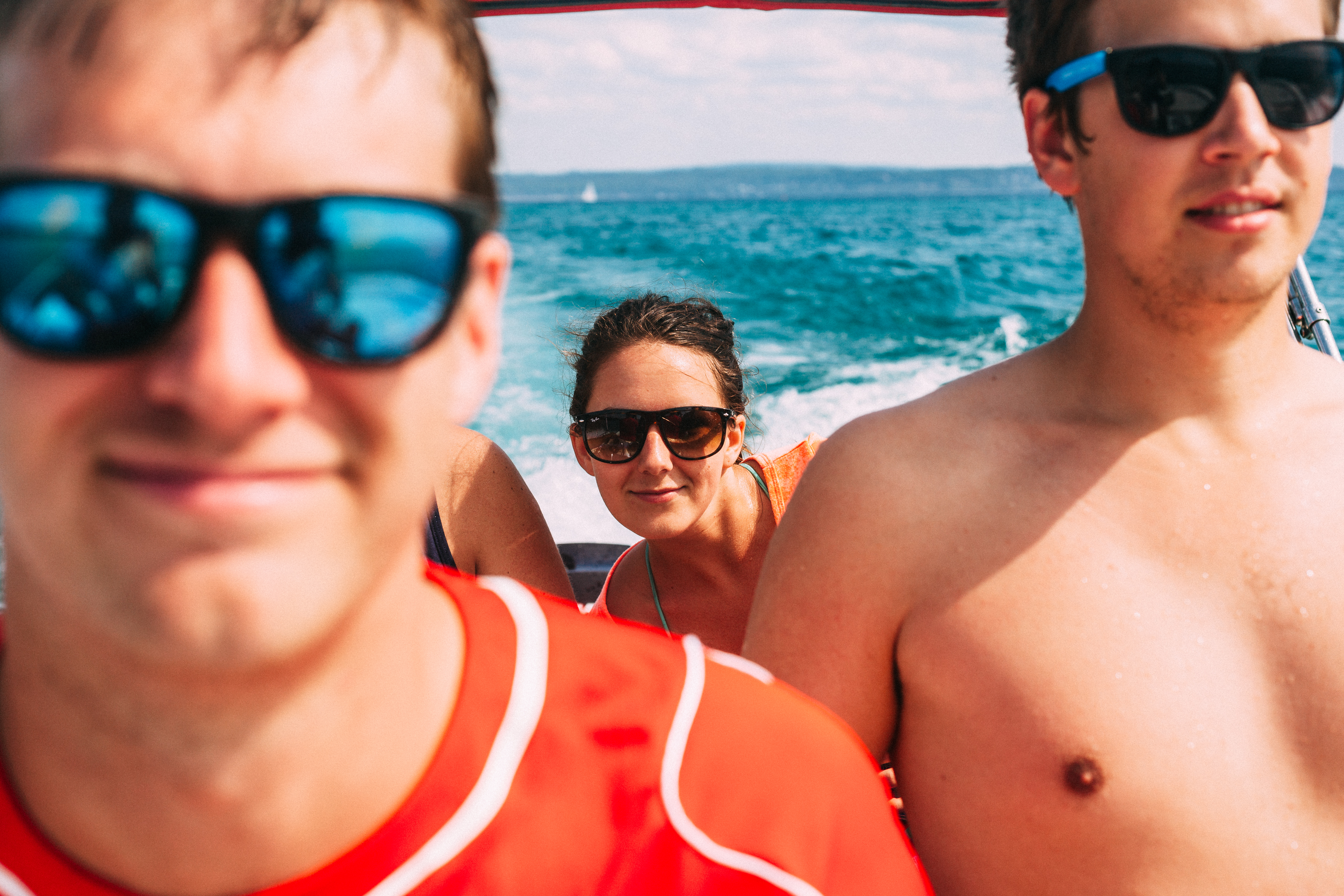 Young woman in dark sunglasses framed between two young men riding in a boat across churning lake water.