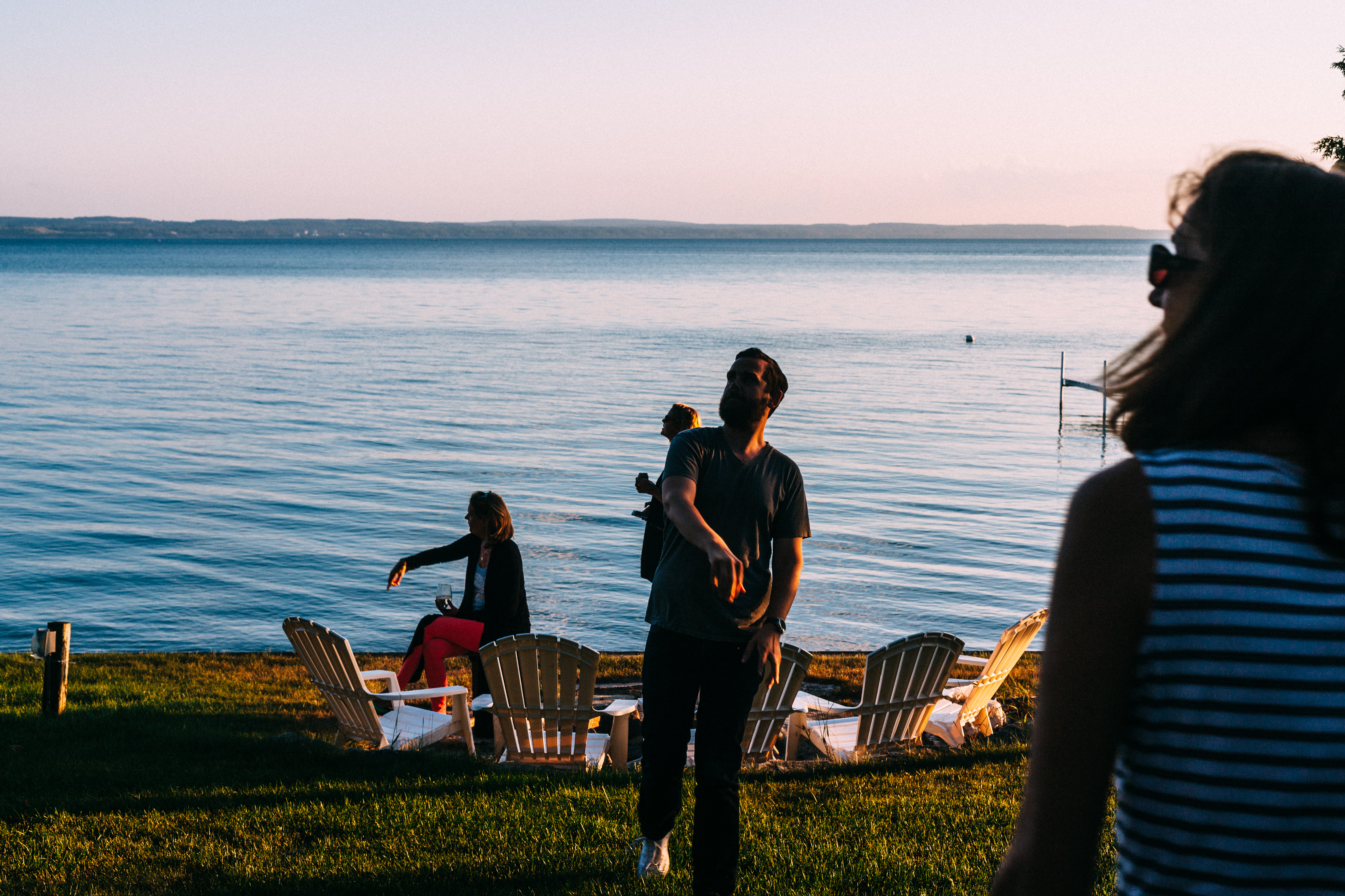 Friends gathered around lawn chairs by a lake at sunset tossing a football in Michigan.
