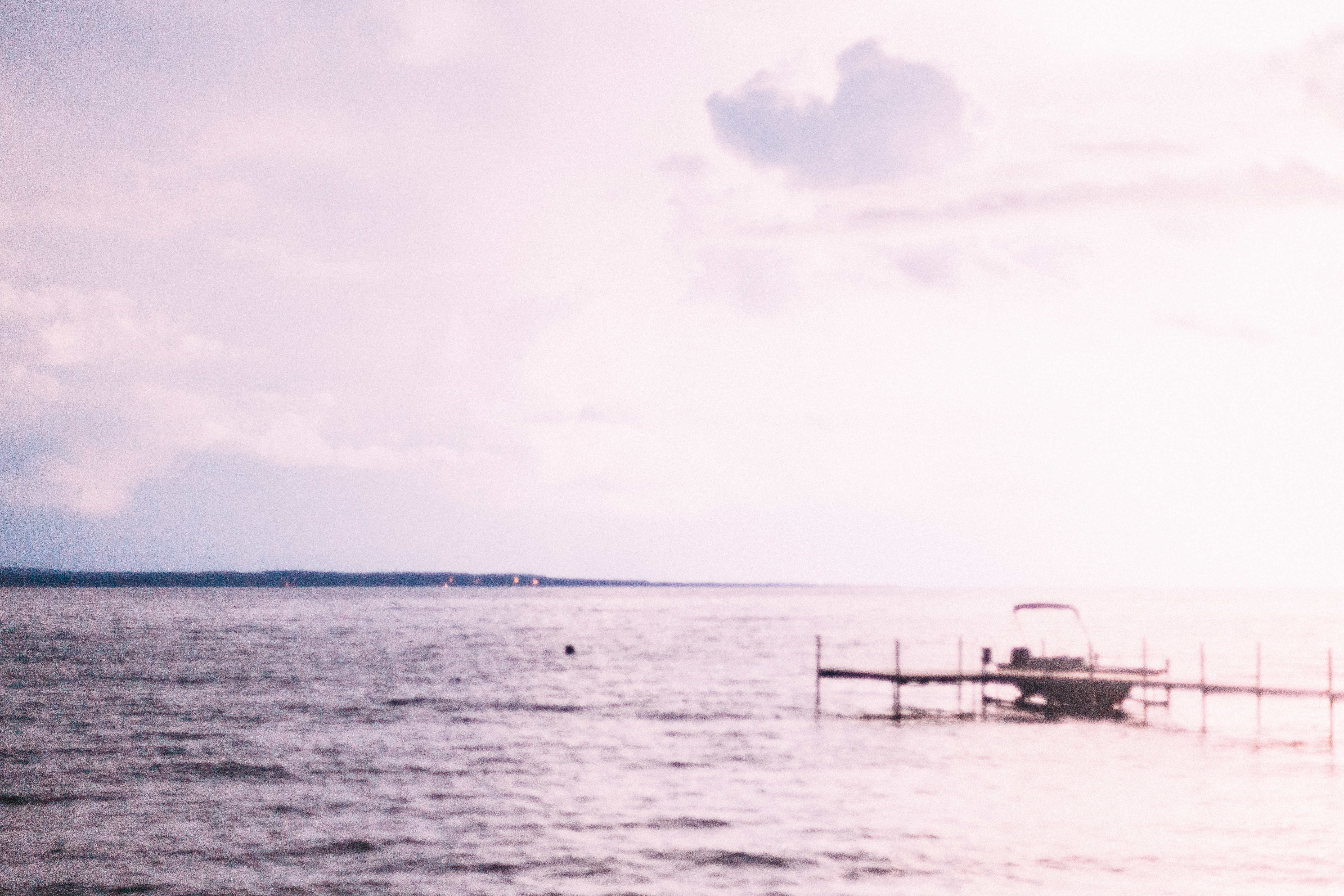 Wooden boat dock and pontoon boat extending into calm lake water under a bright overexposed sky.