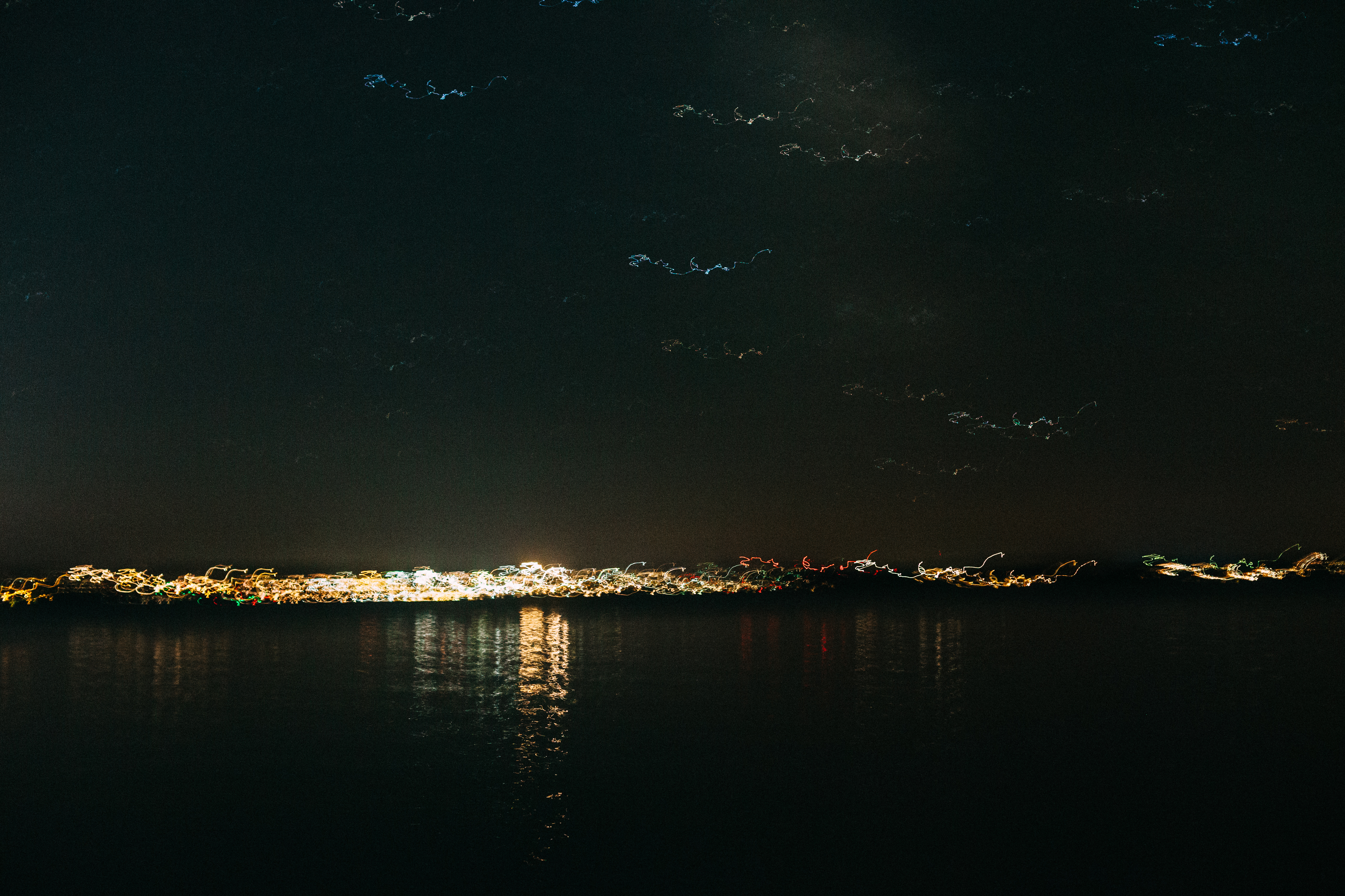 Long exposure light trails of distant city shoreline lights reflected over dark lake water at night.