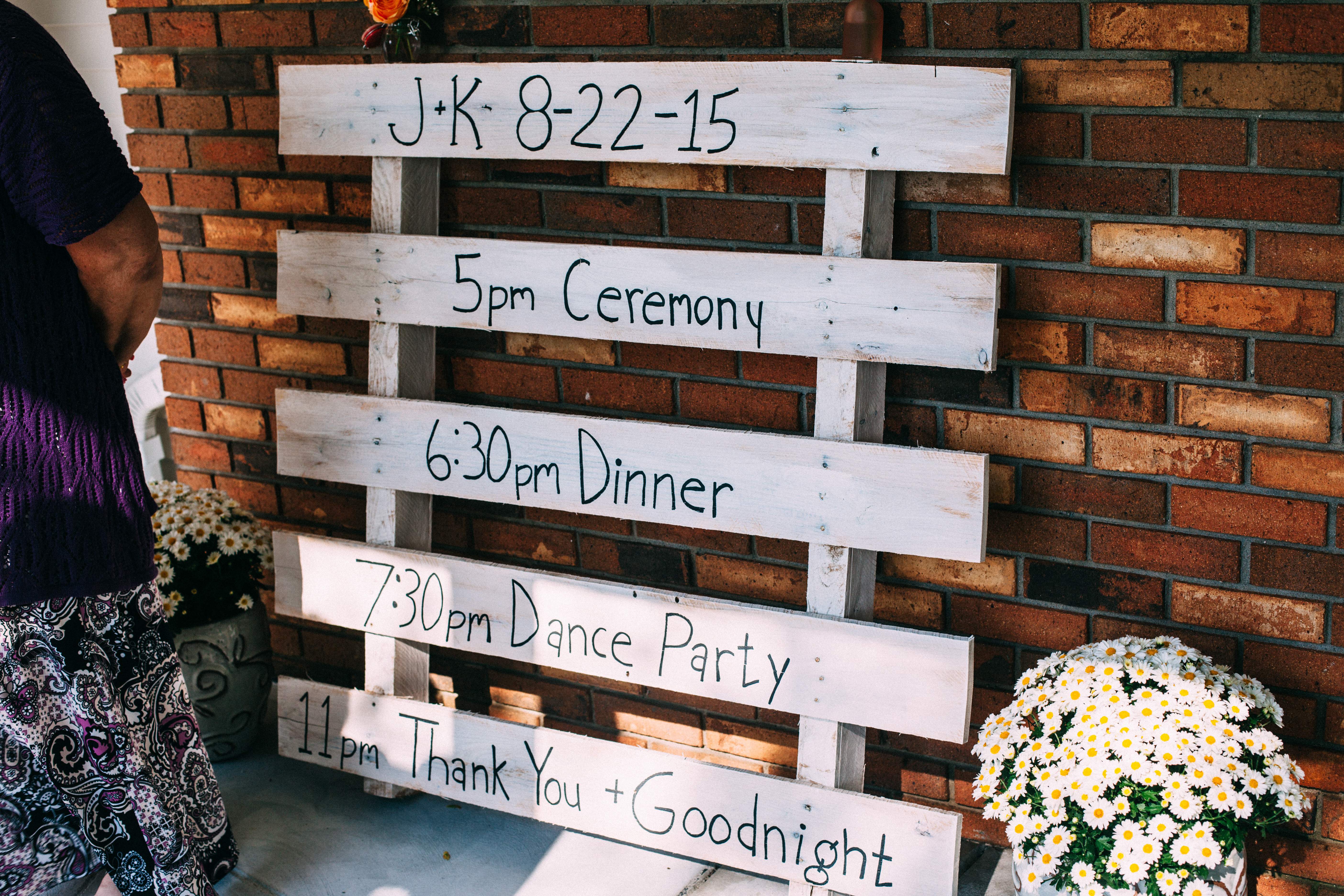 Hand-lettered wooden pallet wedding schedule sign reading 'J+K 8-22-15' flanked by daisy pots.