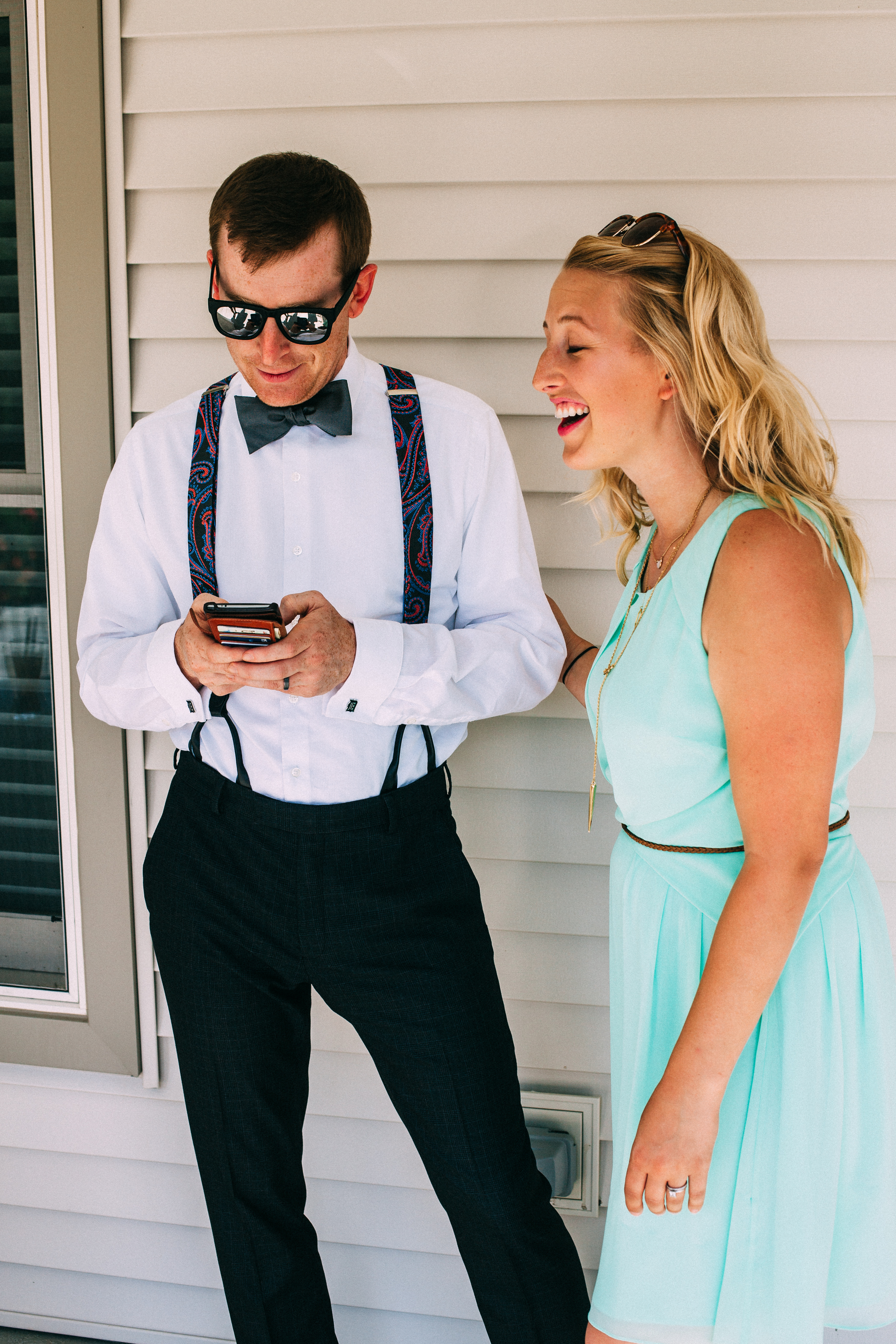 Groomsman in sunglasses, bow tie, and paisley suspenders looking at his phone beside laughing woman in mint dress.