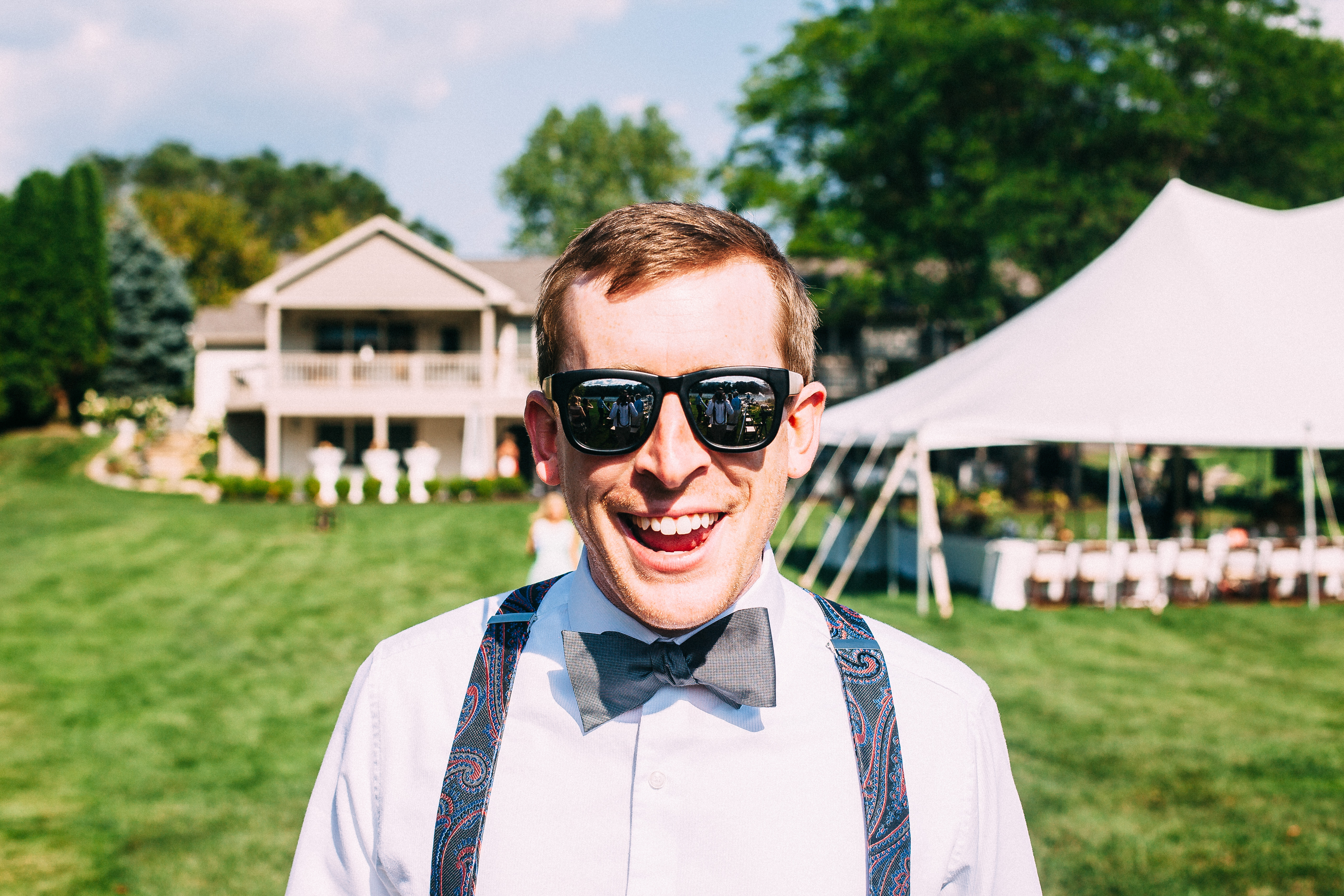 Laughing groomsman wearing sunglasses, bow tie, and paisley suspenders before a wedding lawn and tent.