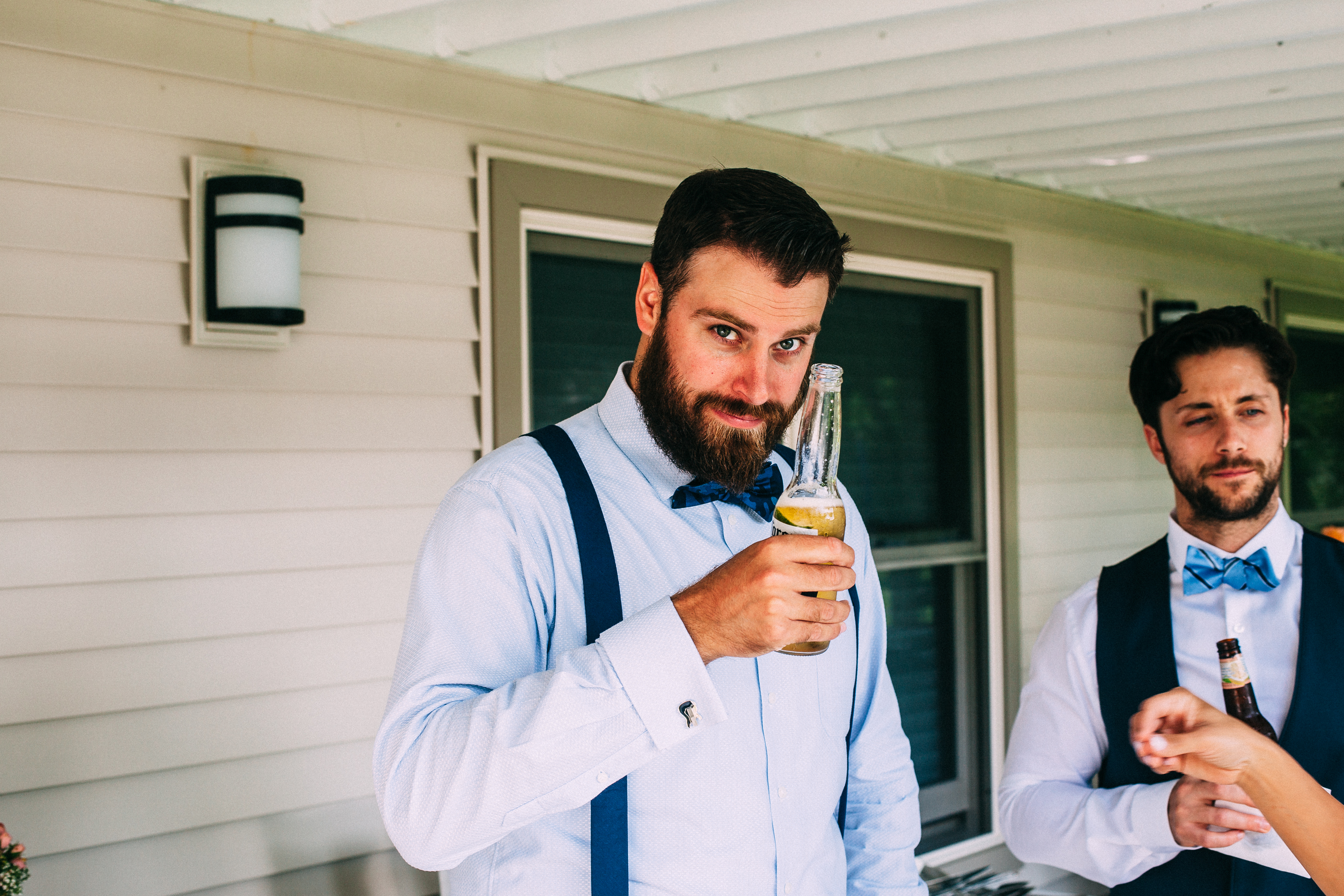 Bearded groomsman in blue shirt and bowtie smiling while holding a beer bottle with a lime wedge on a porch.