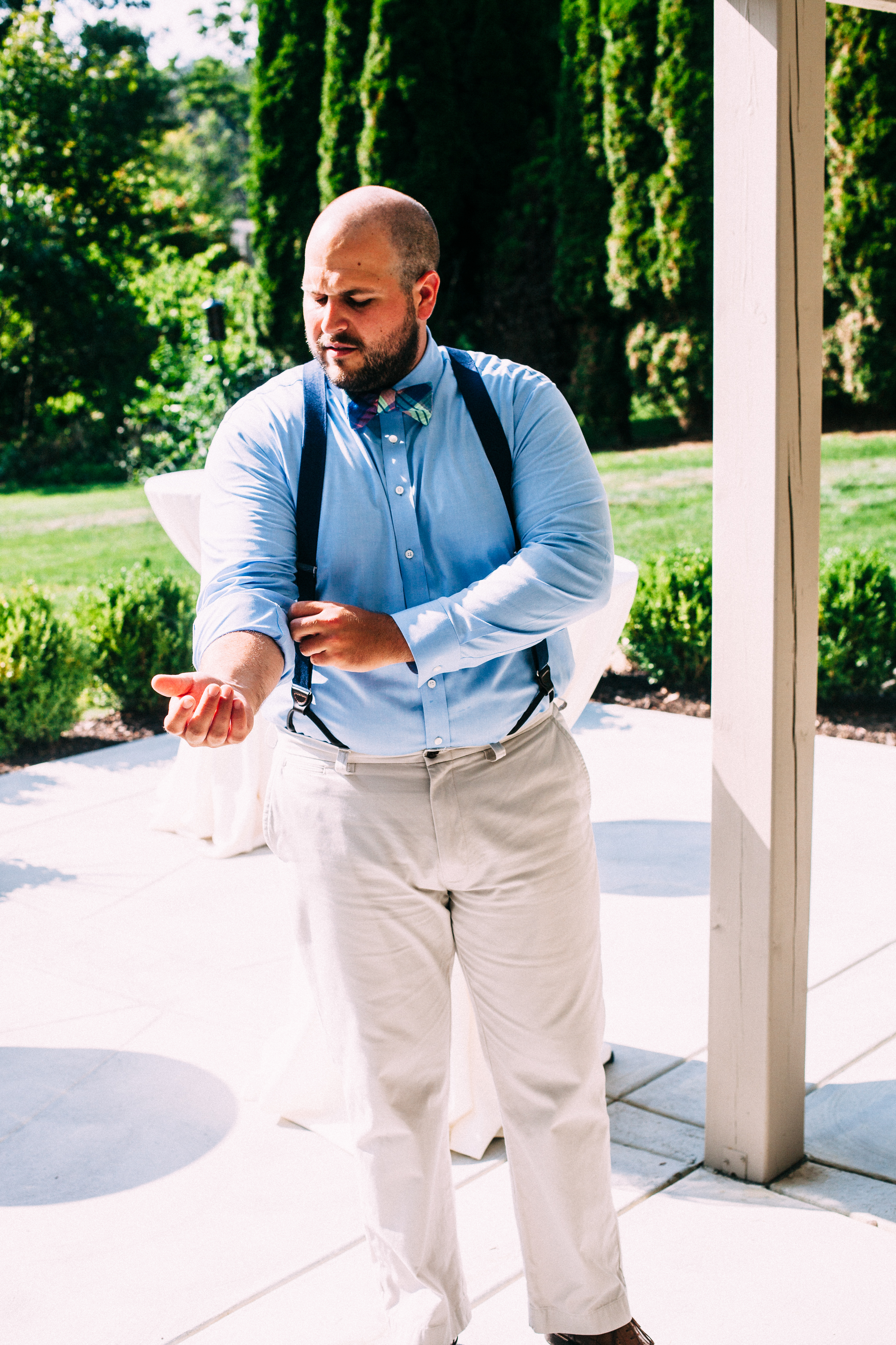 Bearded groomsman in suspenders and plaid bow tie adjusting his shirtsleeve on a sunny outdoor patio.