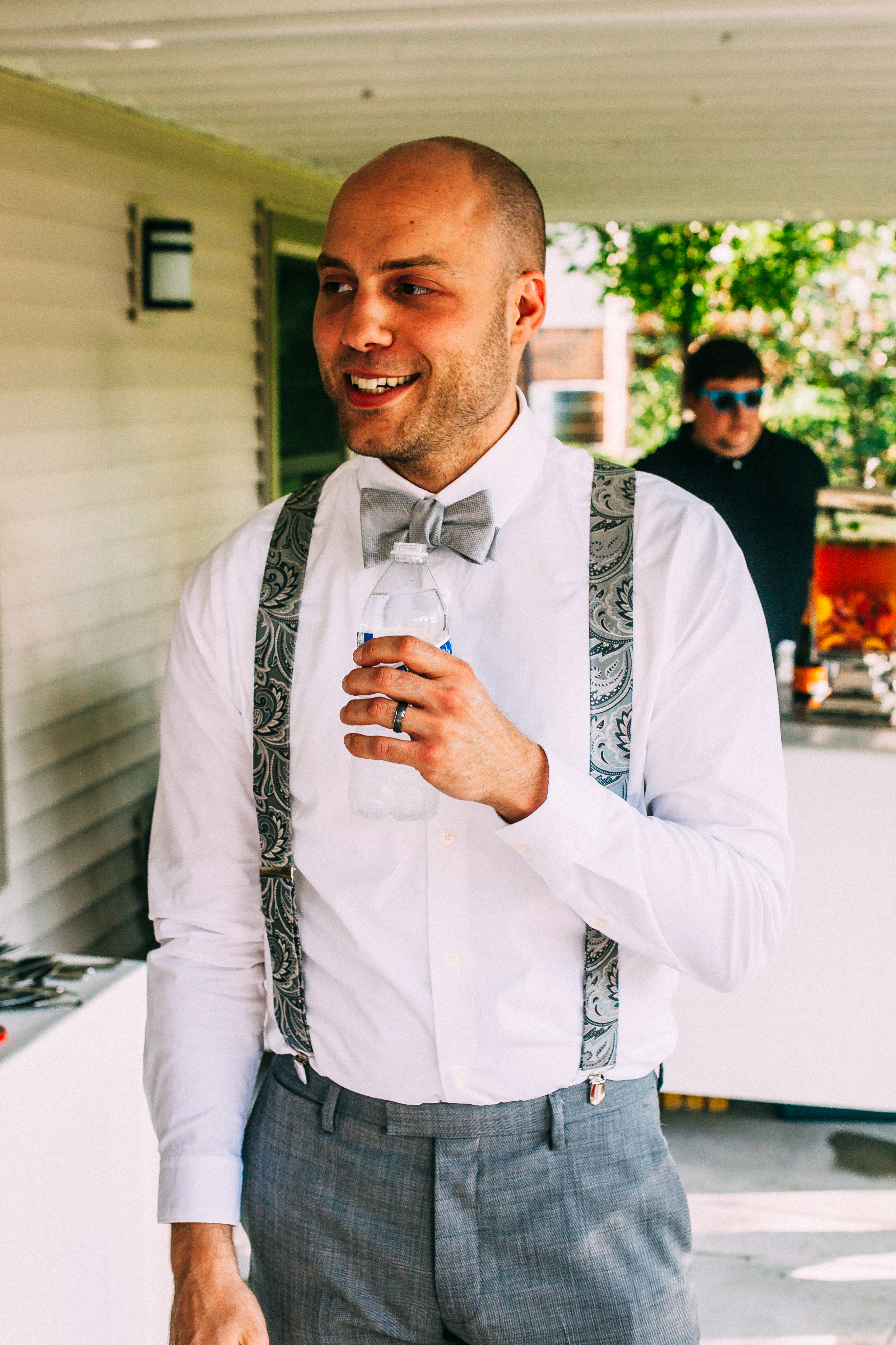 Smiling groomsman with shaved head holding a water bottle in grey bow tie and paisley suspenders.